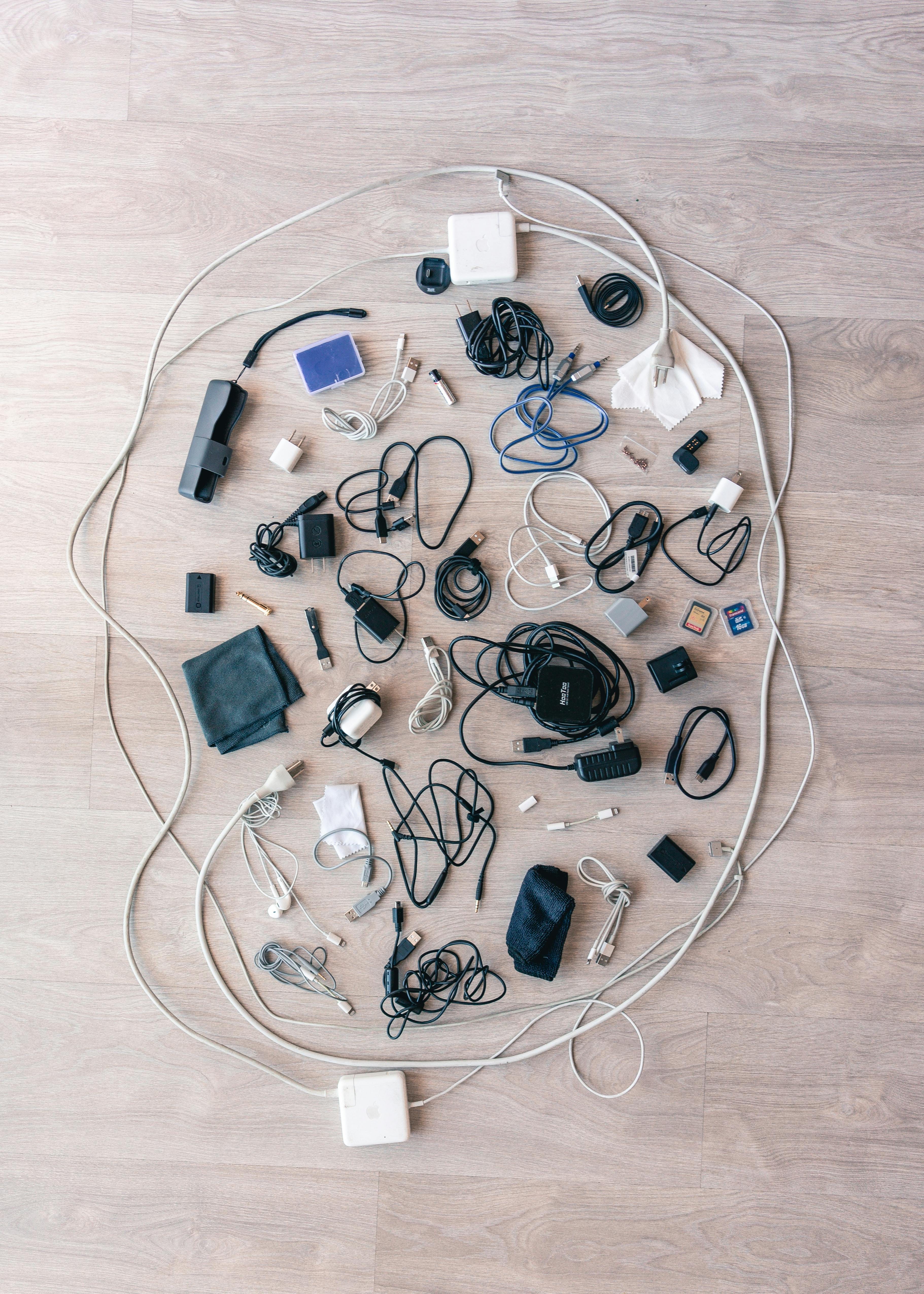 Various cables, chargers, adapters, SD cards, and small electronics spread out on a light wood floor.