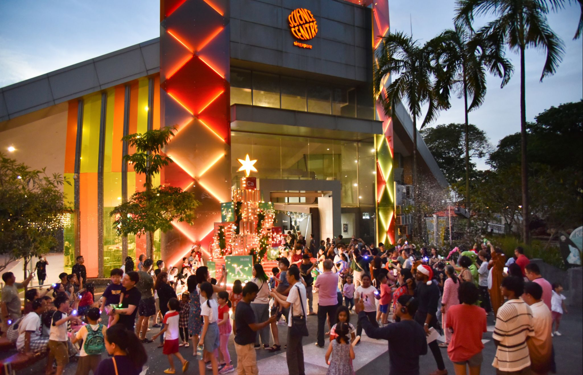 A large crowd gathers outside Science Centre Singapore around a lit Christmas tree with a star topper at dusk.