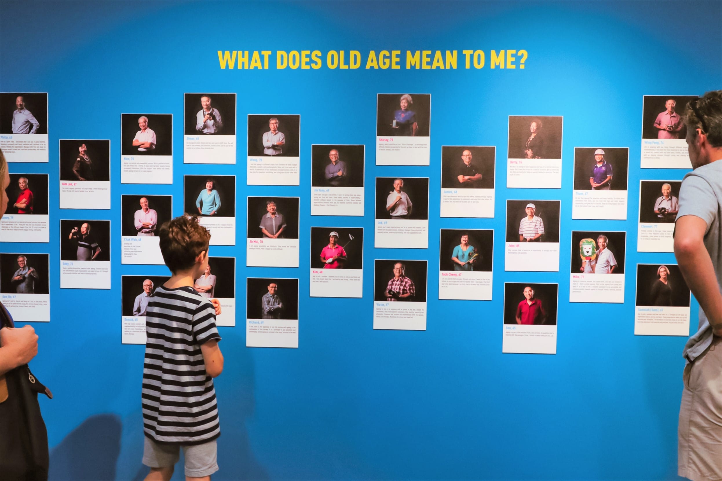 Blue museum wall displays "What Does Old Age Mean To Me?" with many portrait photos, viewed by a child and adults.