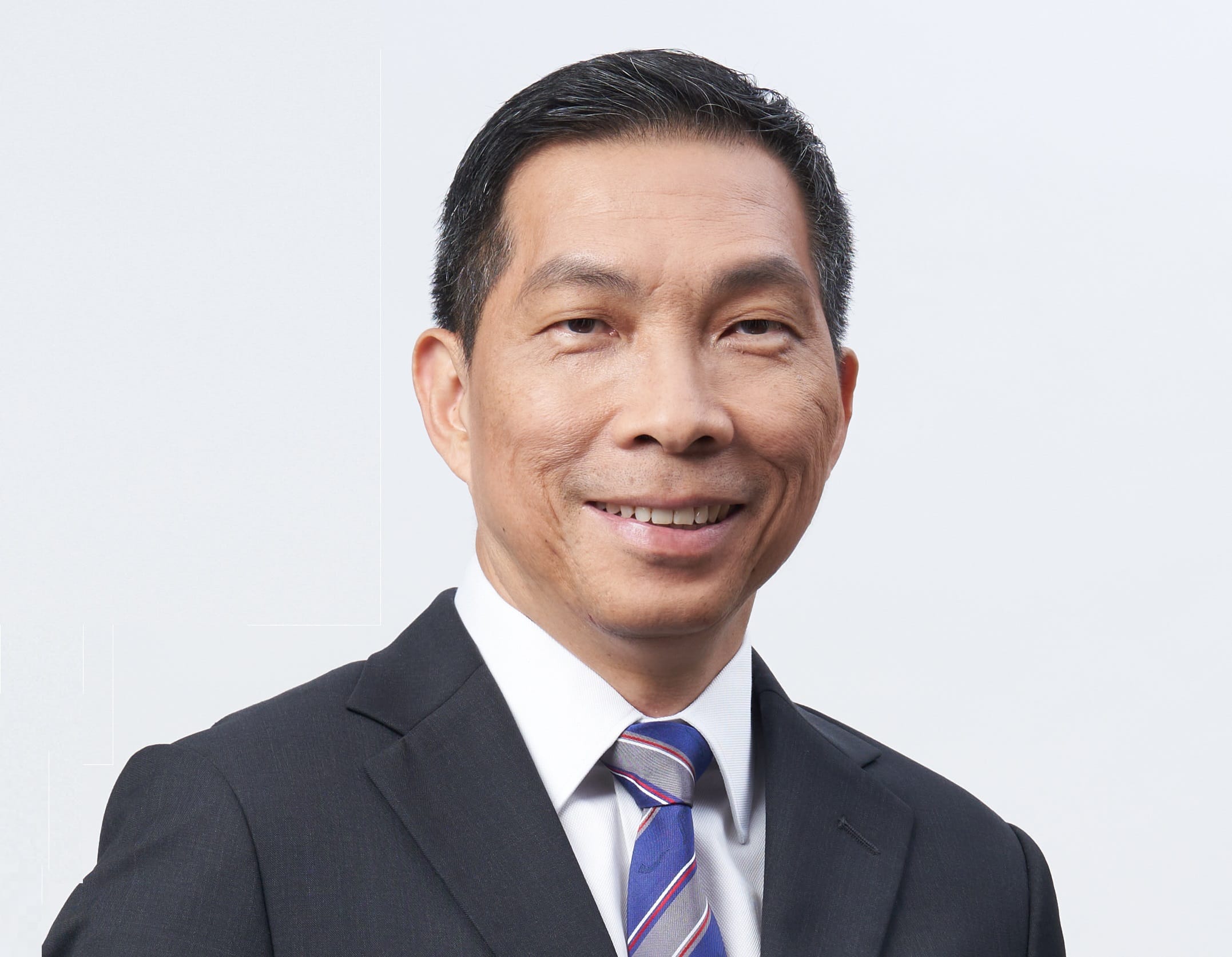 A smiling middle-aged man in a dark suit, white shirt, and blue striped tie against a light gray background.