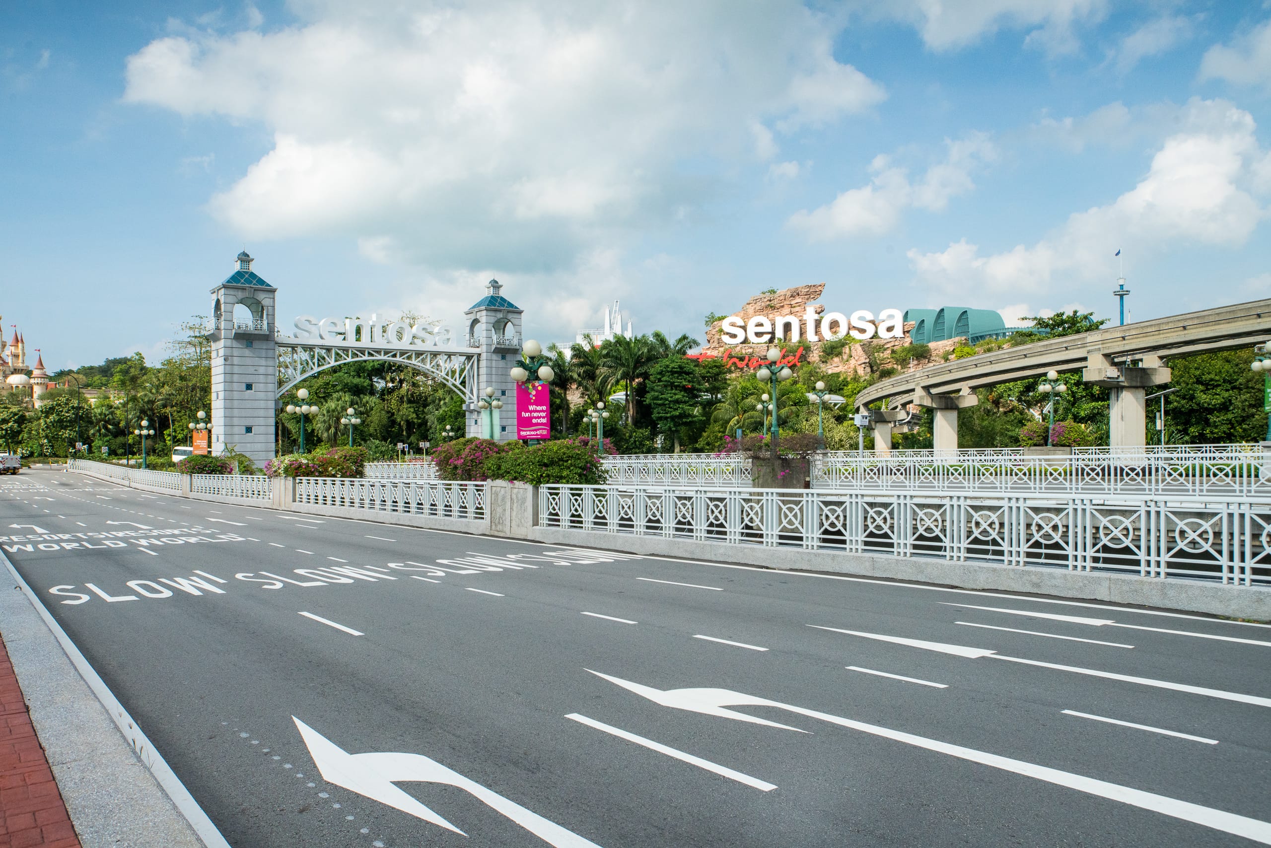 Open road leading to Sentosa island entrance gate with arch, signs, lush greenery, and an elevated rail track.