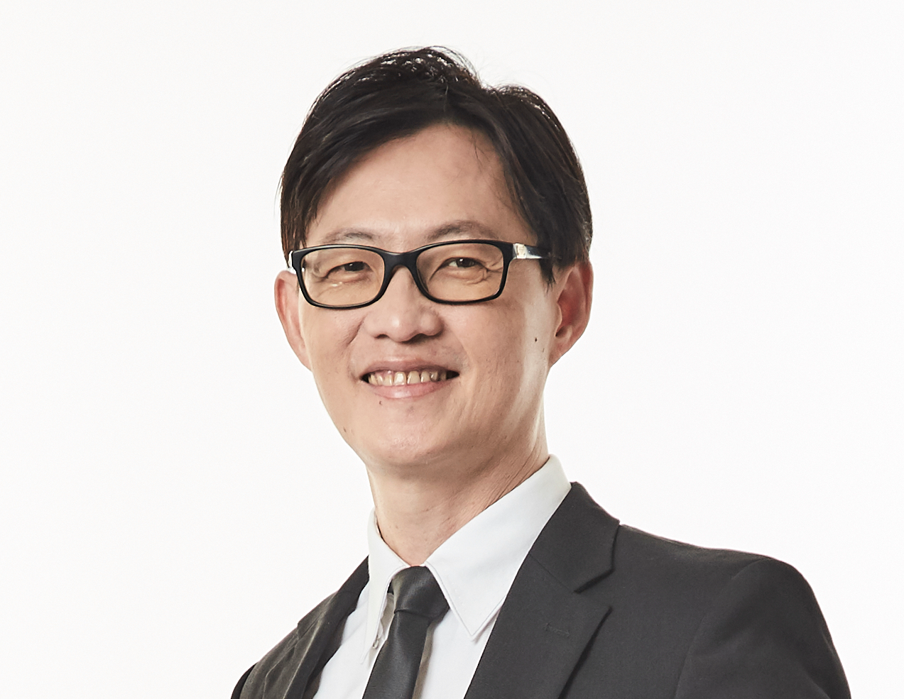 A smiling middle-aged man wearing black-framed glasses, a dark suit, white shirt, and dark tie against a white background.