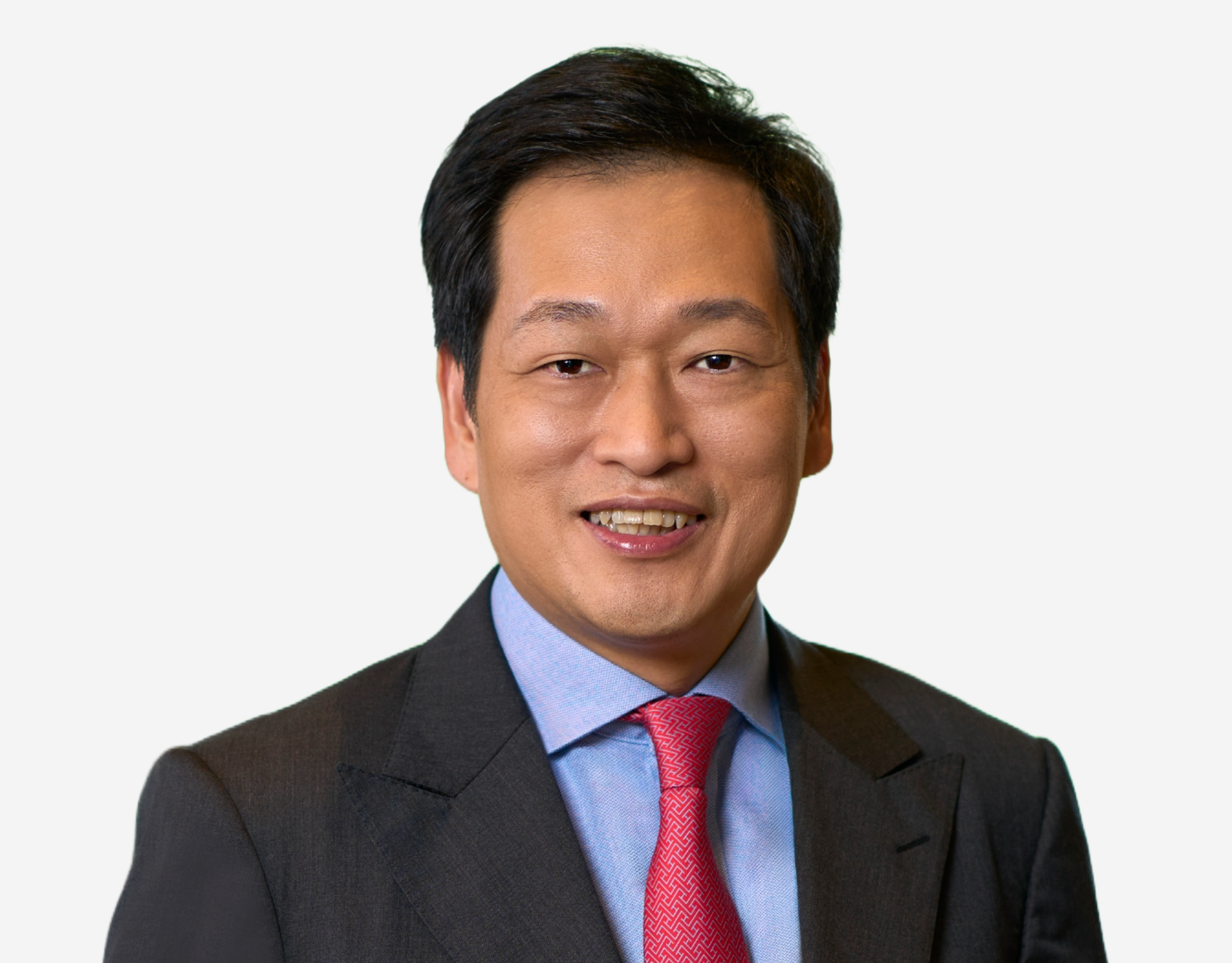 A smiling middle-aged man in a dark suit, light blue shirt, and red patterned tie against a gray background.