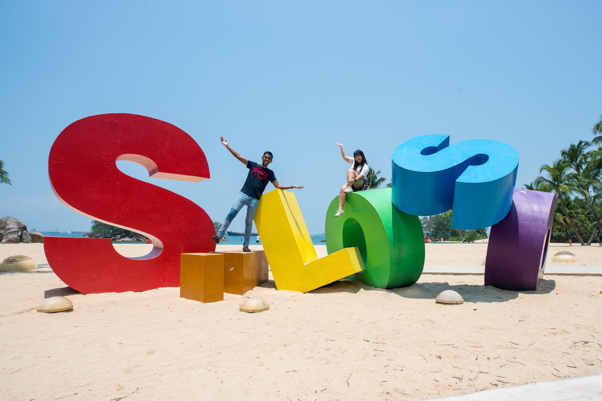 Two people pose on large colorful letter sculptures spelling Siloso on a sunny sandy beach with palm trees.