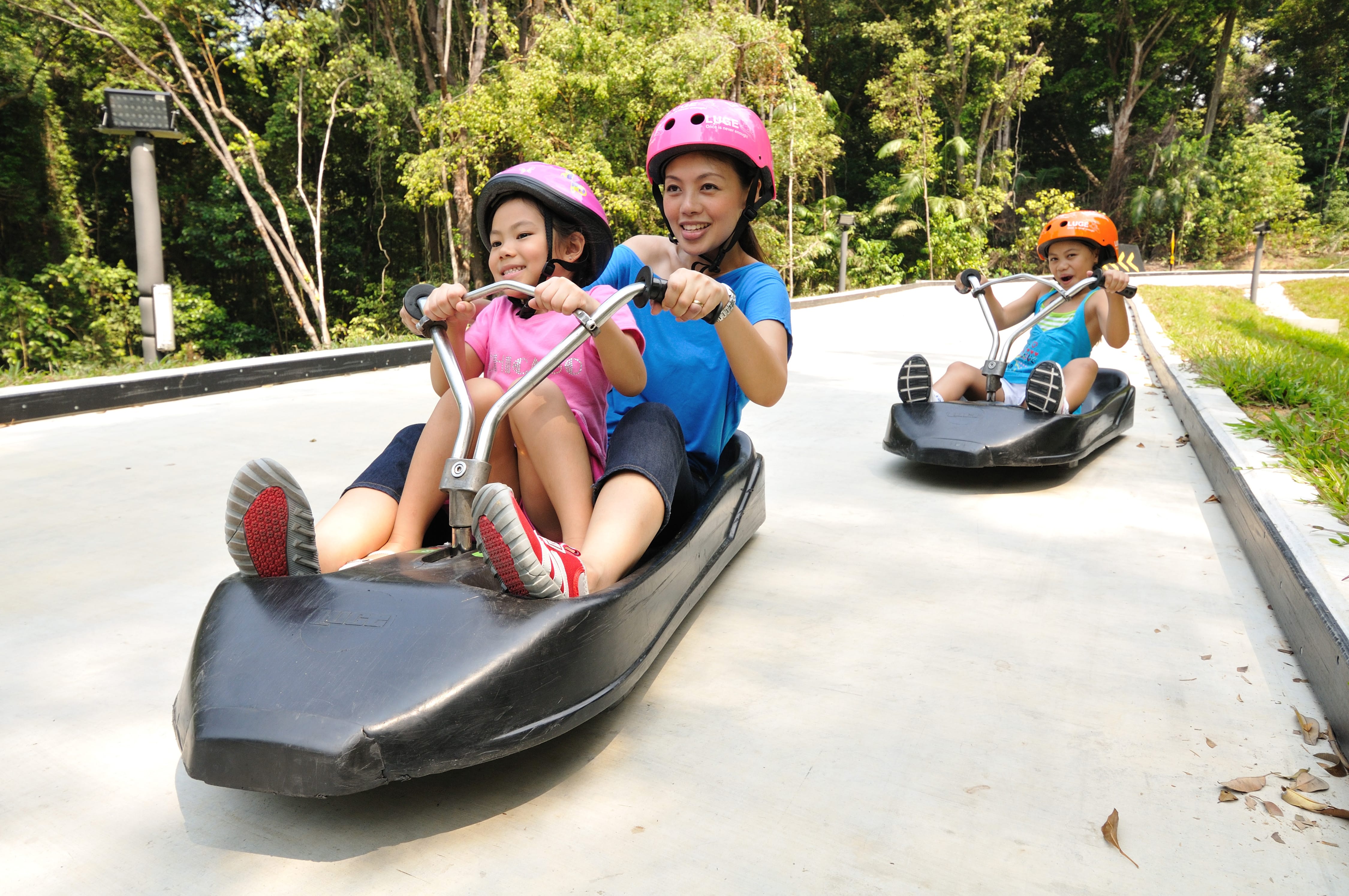 Three people wearing helmets ride luge carts down an outdoor concrete track surrounded by green trees.