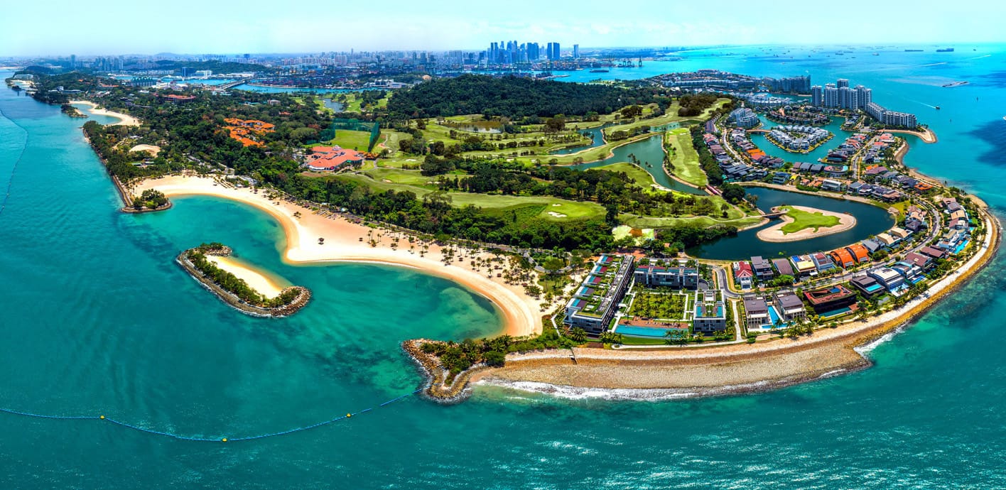 Aerial view of a lush island with sandy beaches, a golf course, waterways, and a city skyline in the background.