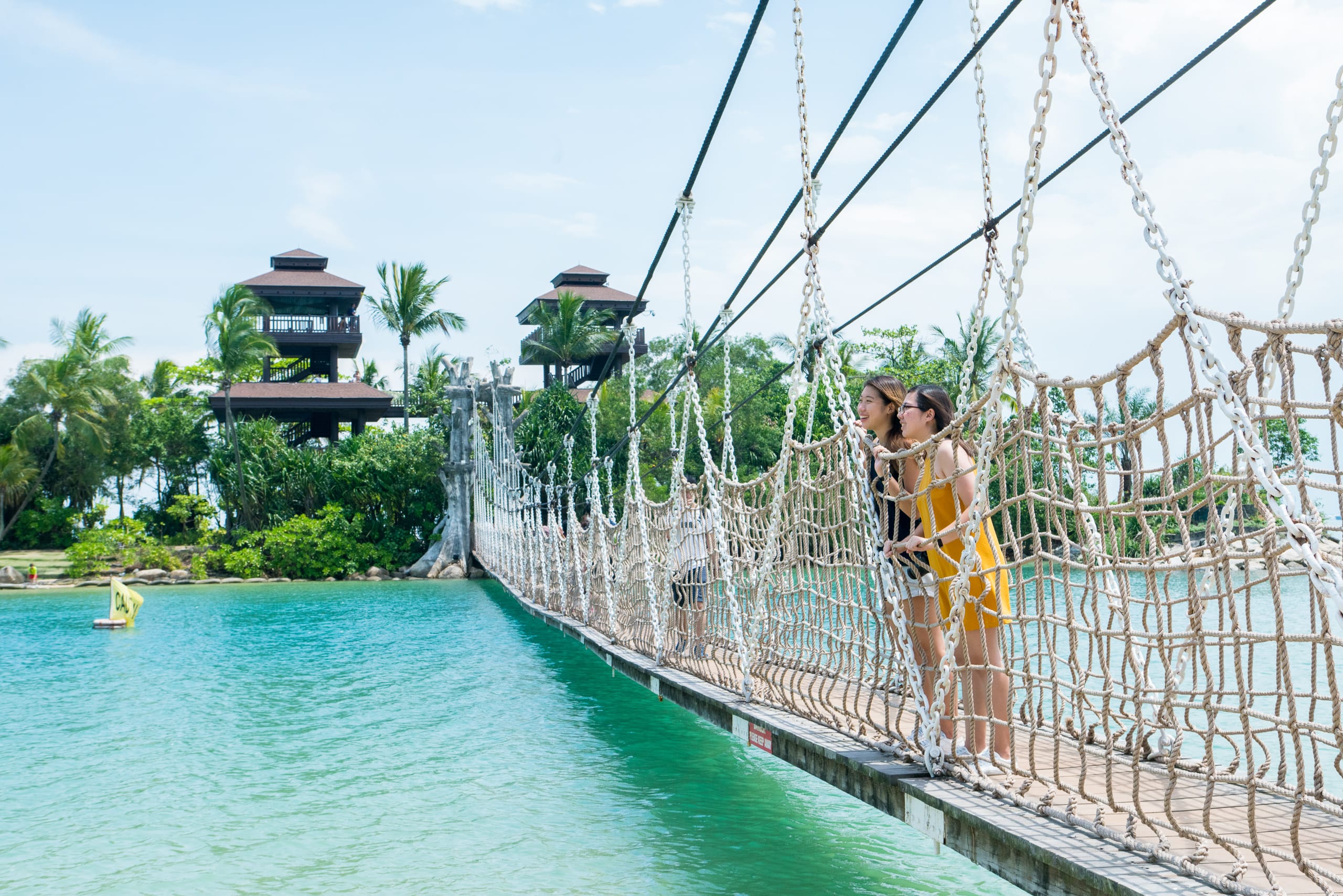 Two women stand on a rope suspension bridge over turquoise water, with tropical trees and wooden towers behind them.