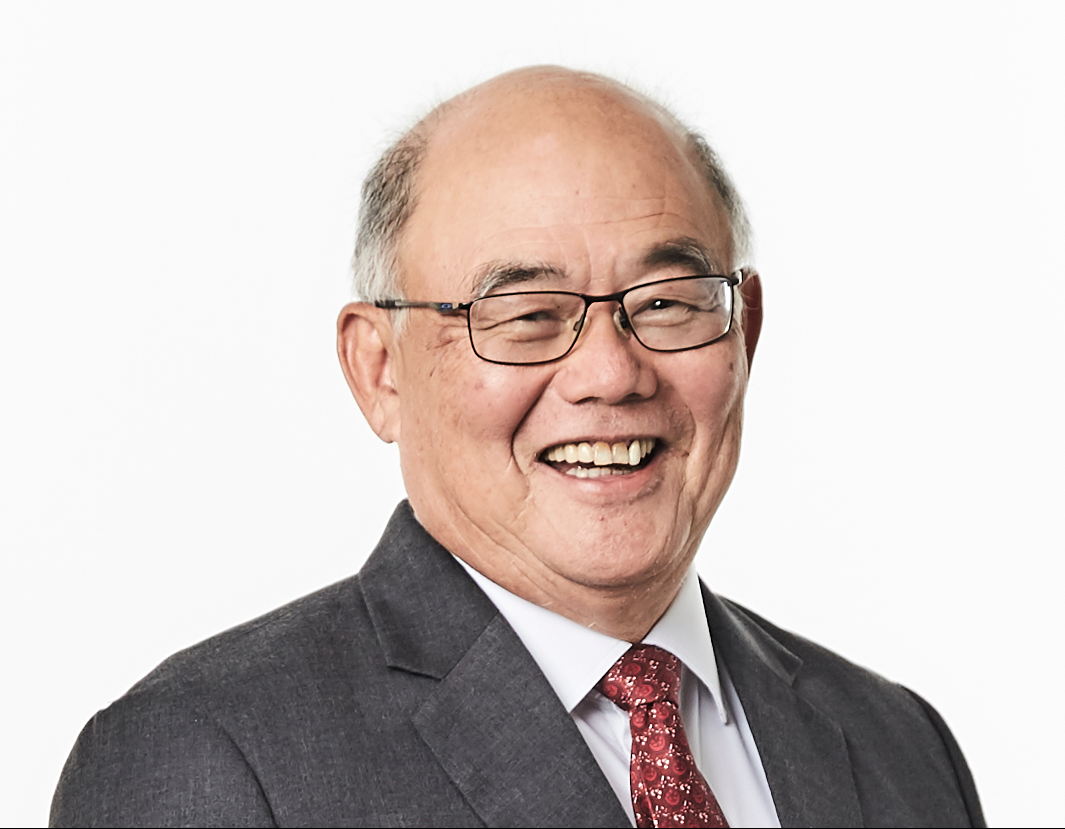 Older man with glasses smiling broadly, wearing a gray suit, white shirt, and red patterned tie against a white background.