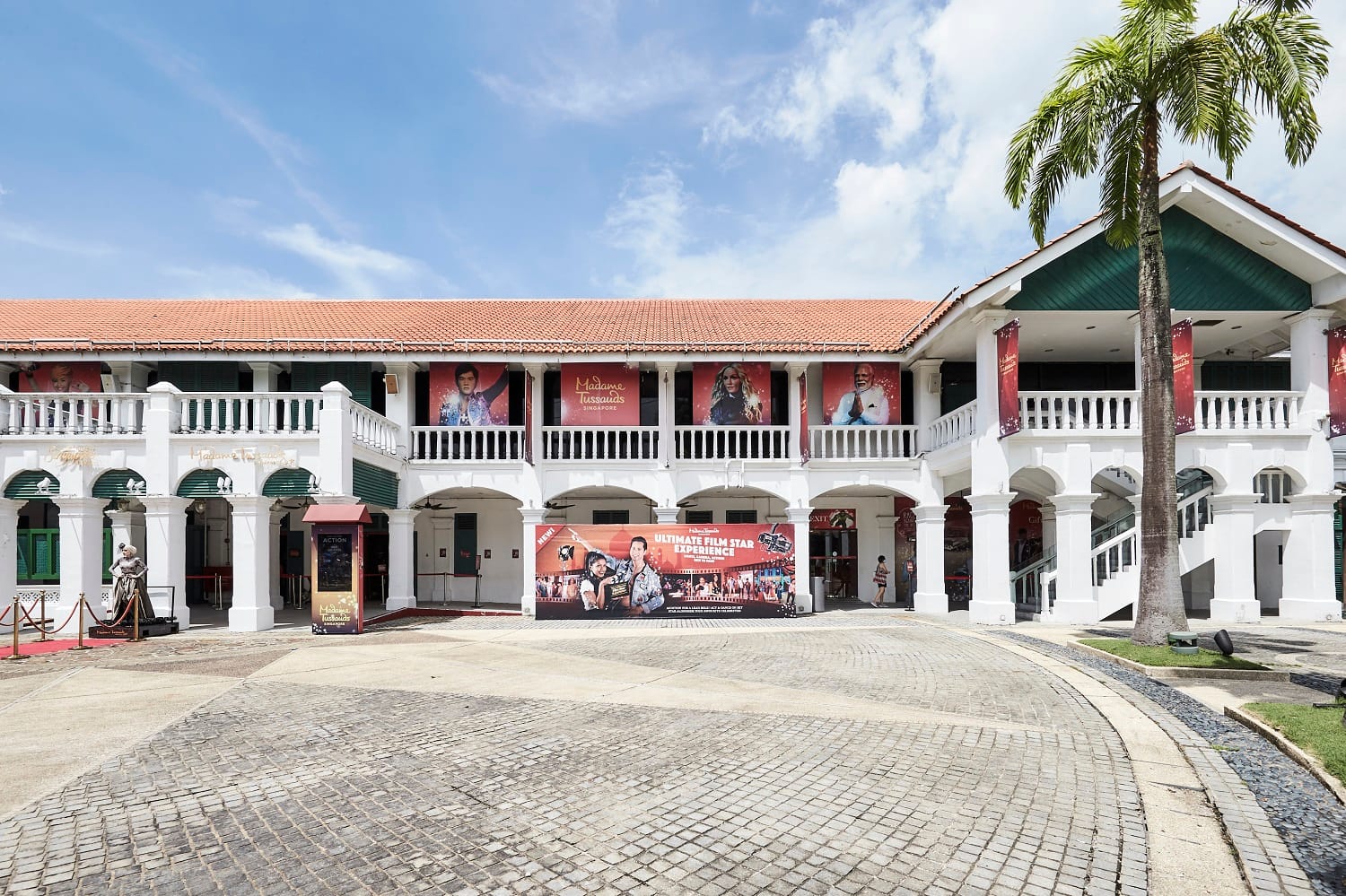 White colonial-style building with red tile roof housing Madame Tussauds Singapore, with celebrity banners and a palm tree.