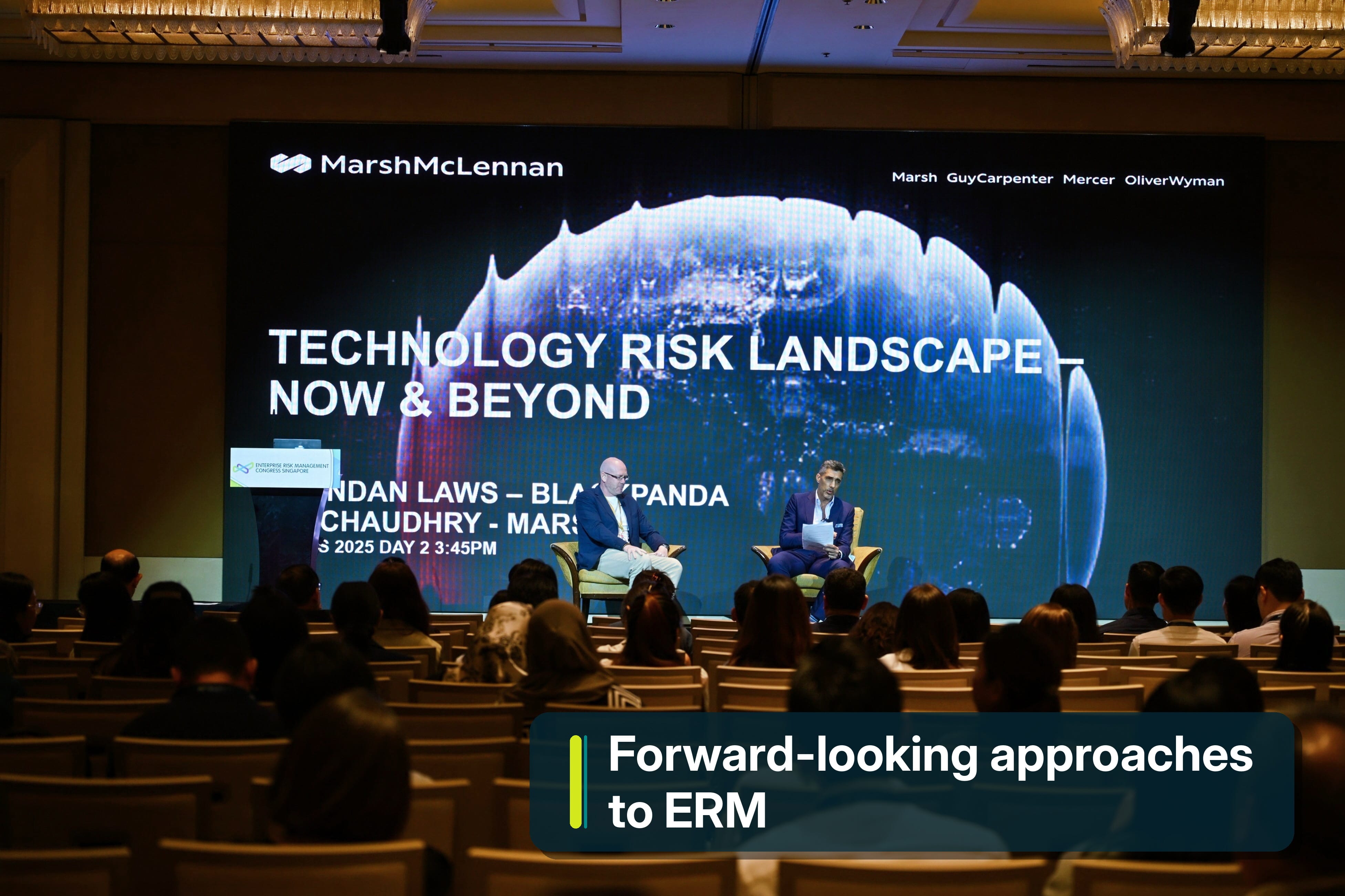 Two male speakers seated on stage before an audience, having a discussion.