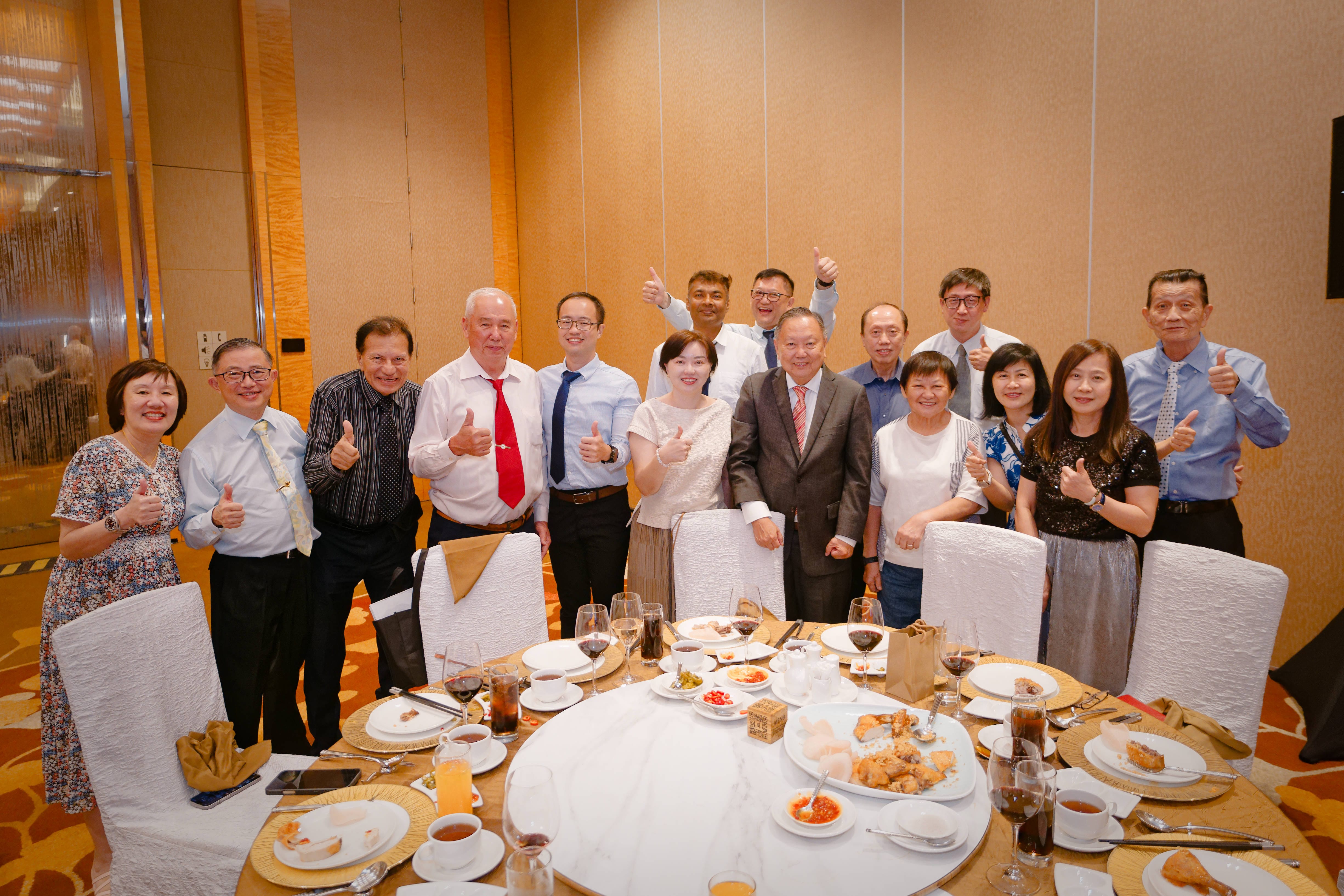 A large group of banquet guests stand together around a fully set round dining table, smiling and giving thumbs-up gestures for the camera.