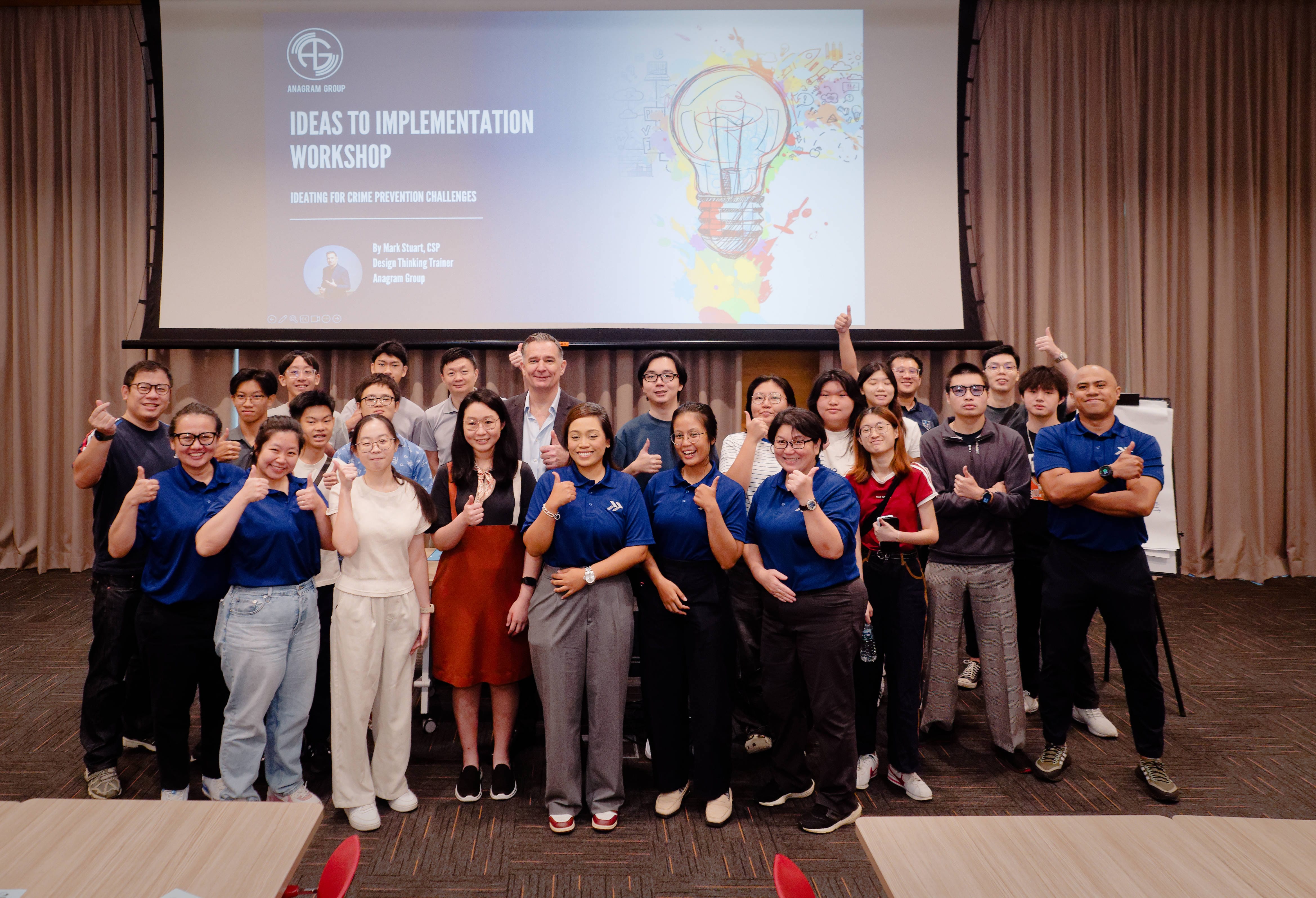 A wide-angle group photo captures diverse participants and facilitators smiling and giving thumbs-up gestures inside a function room during the "Ideas to Implementation Workshop" hosted by Anagram Group. A large projector screen behind them displays the workshop title, "Ideating for Crime Prevention Challenges," along with presenter details for Mark Stuart and a colorful graphic of a lightbulb.