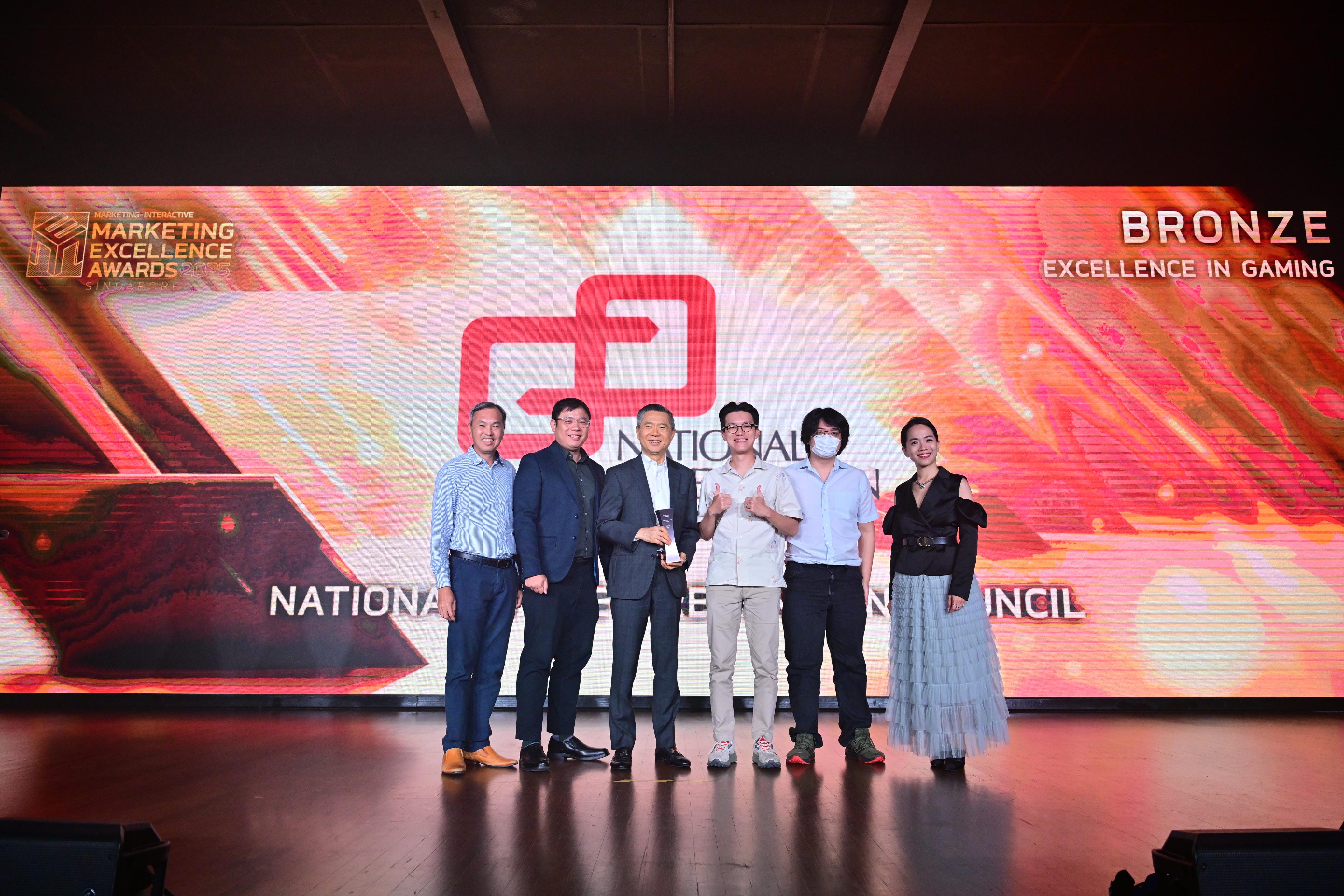 Six people pose on stage at the Marketing Excellence Awards, holding a trophy, with a screen showing Bronze in Gaming.