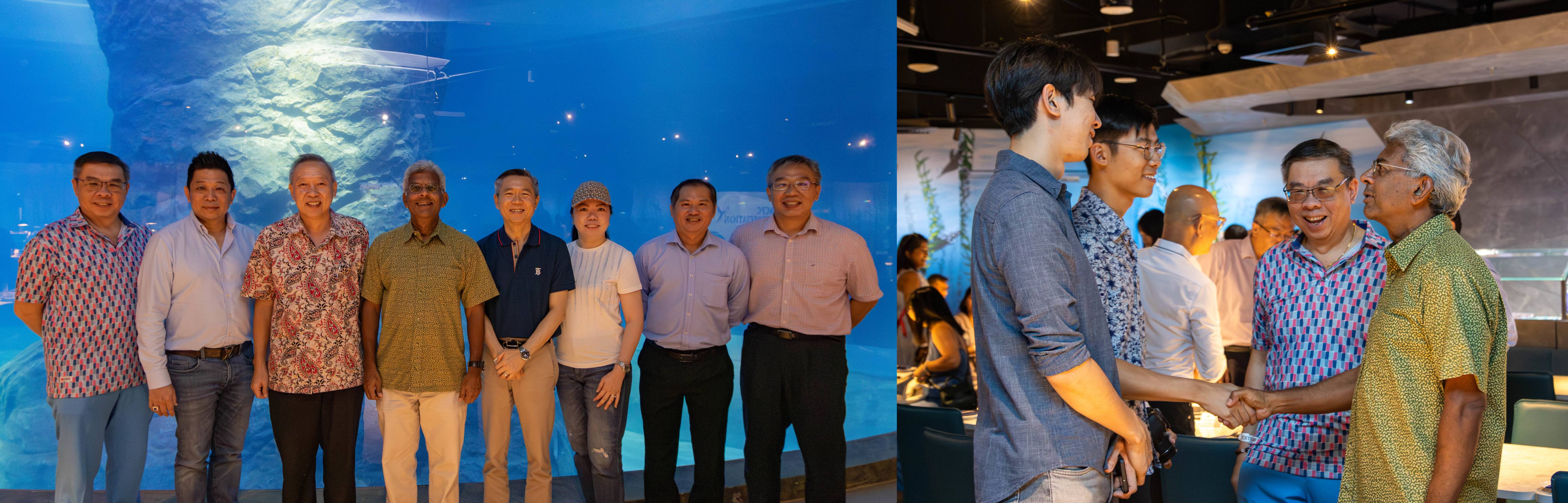 Eight men pose before a blue aquarium tank; beside it, two men shake hands while others converse nearby.