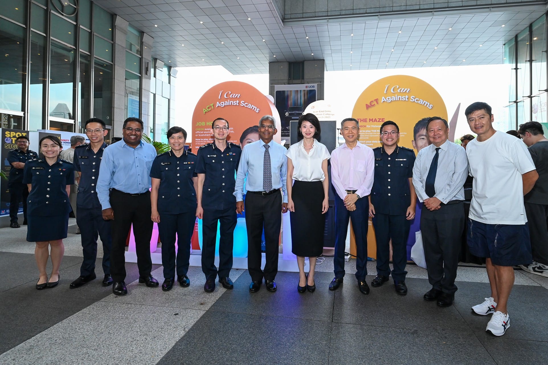 Group of people posing in front of two colorful "I Can ACT Against Scams" campaign banners at an outdoor event.