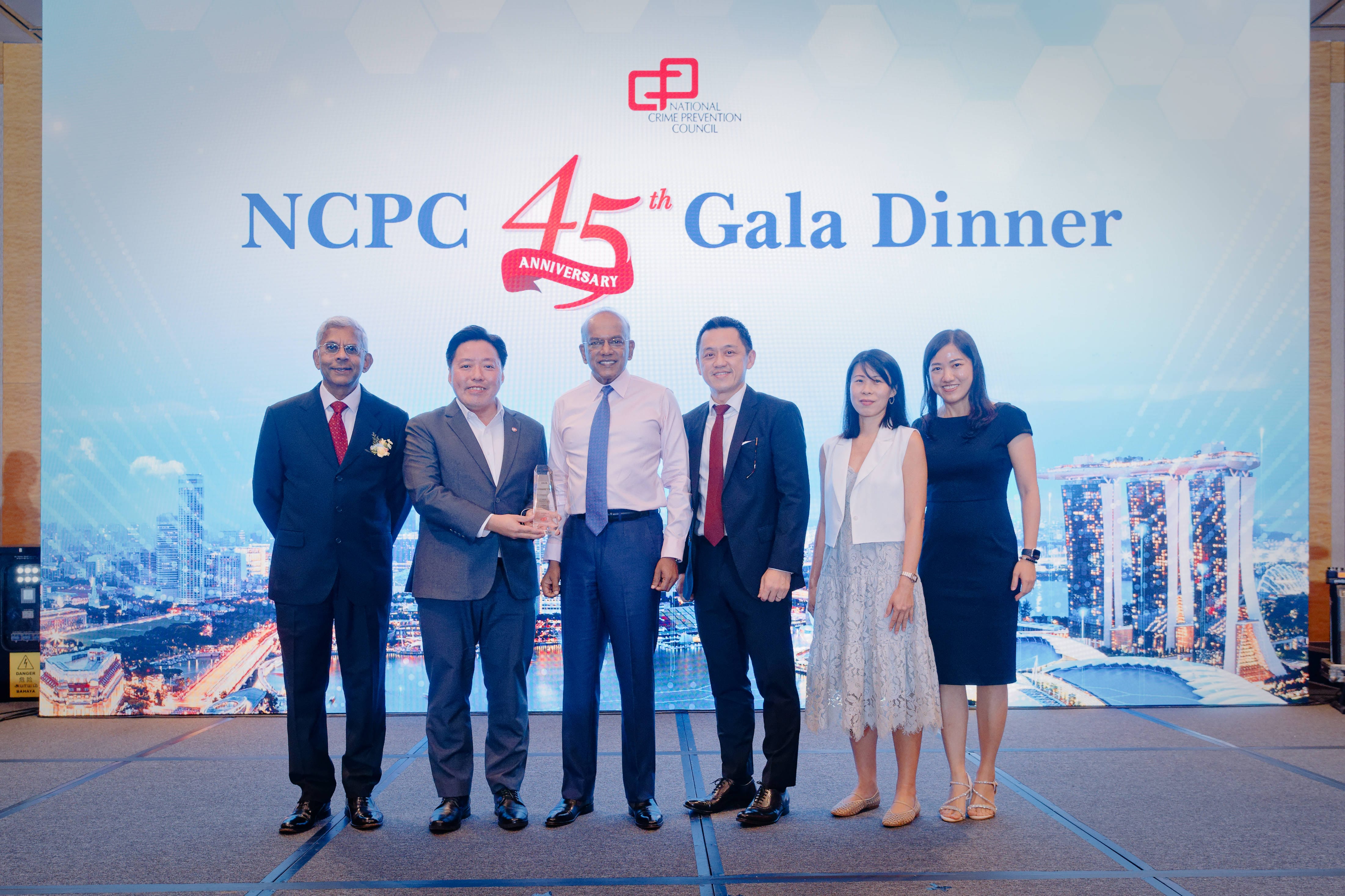 A formal stage presentation captures a group of six individuals posing on stage in front of the NCPC 45th Gala Dinner backdrop, with two men at the center presenting and receiving a glass award trophy.