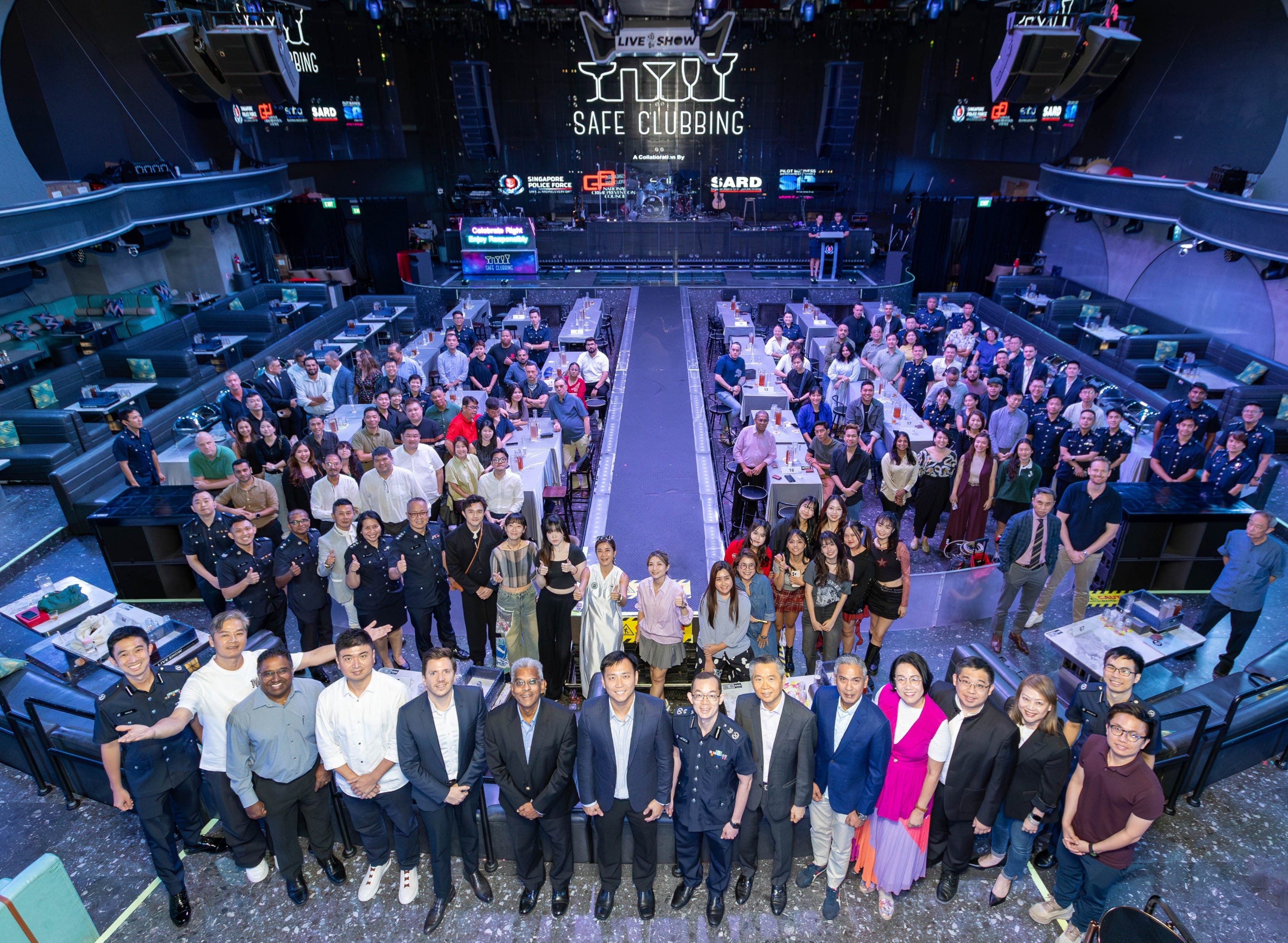 Large group posing in a nightclub venue under a lit "Safe Clubbing" sign with tables and a stage behind them.