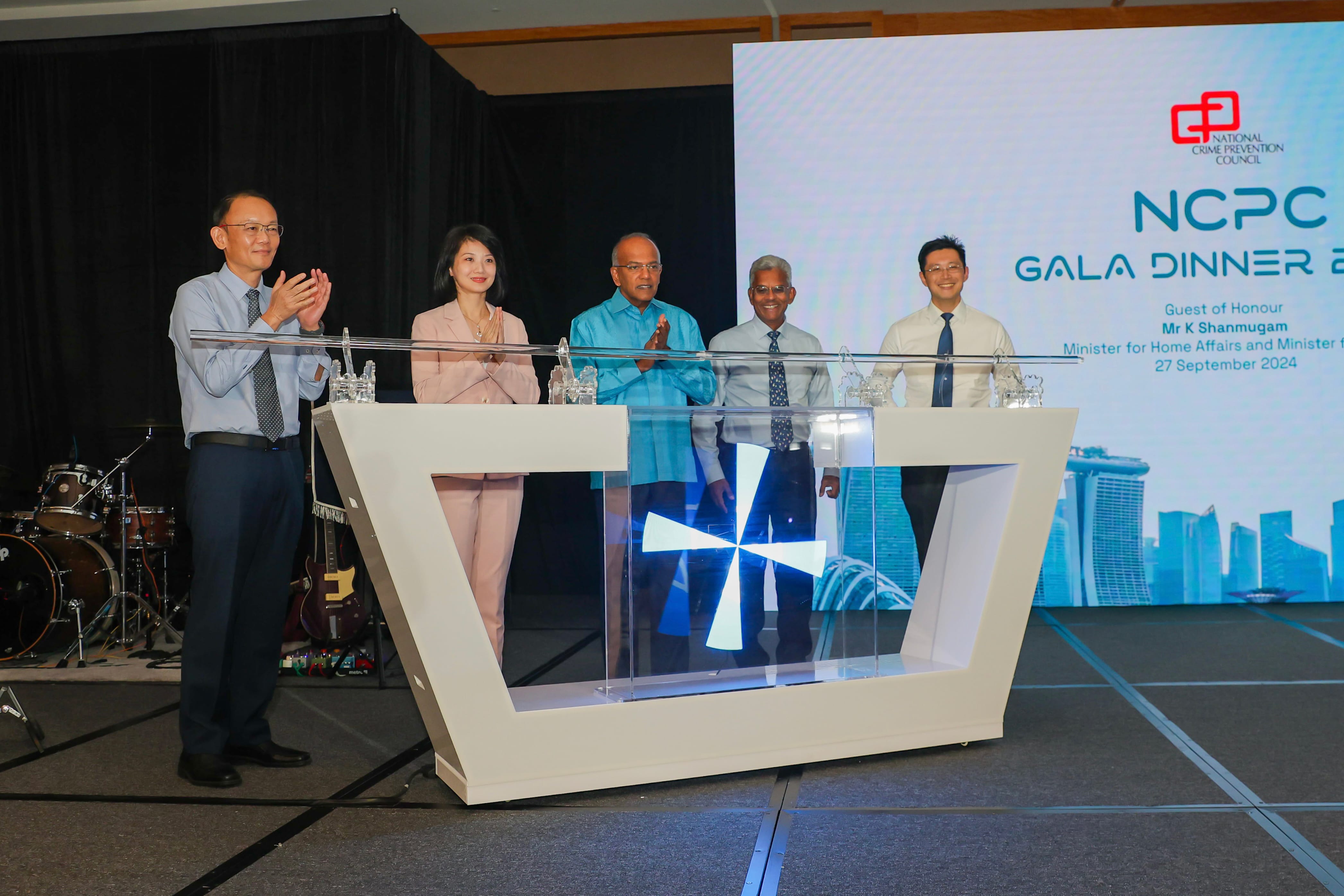Five people stand on stage at the NCPC Gala Dinner 2024, clapping near a lit display podium.