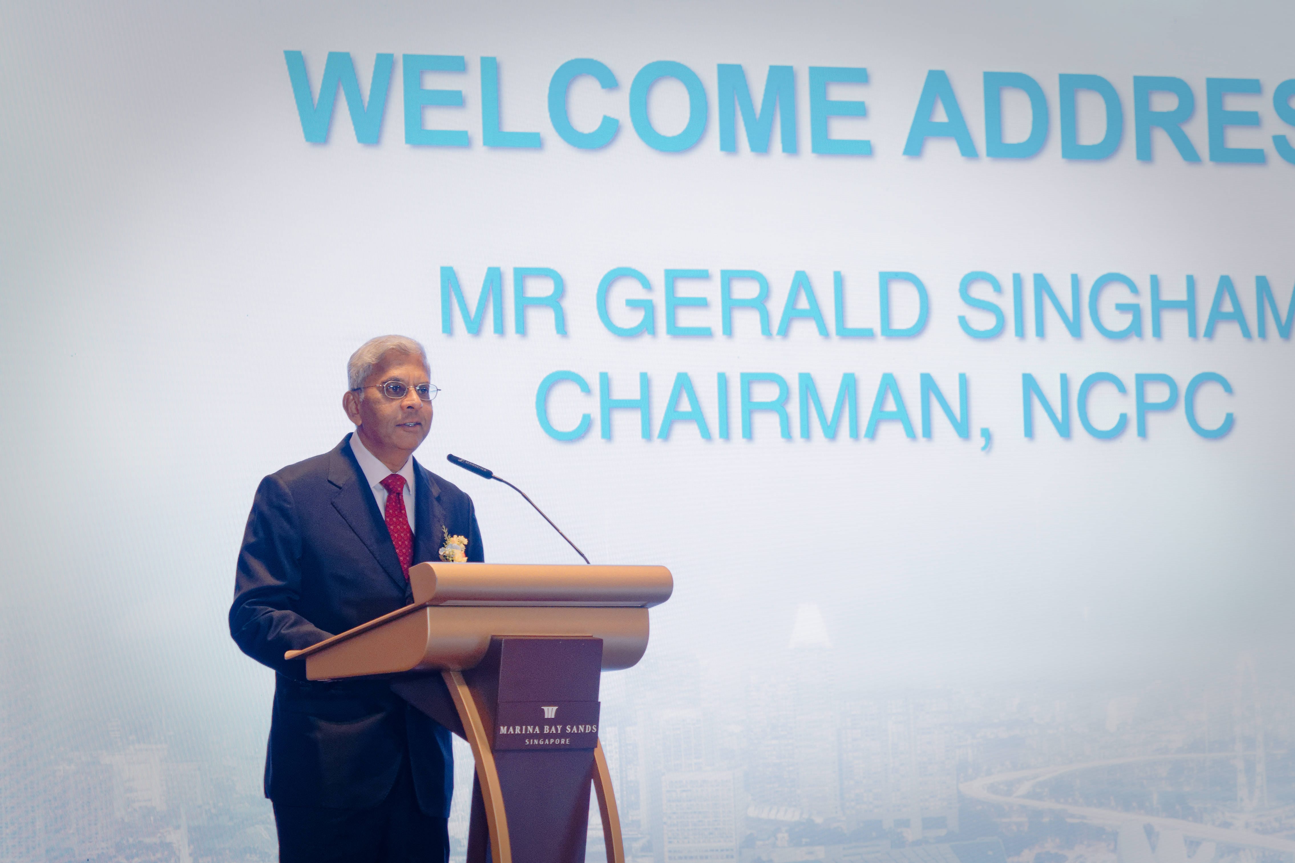 A formal individual portrait of Mr. Gerald Singham, Chairman of the NCPC, delivering a welcome address at a wooden podium with the main projection screen behind him.