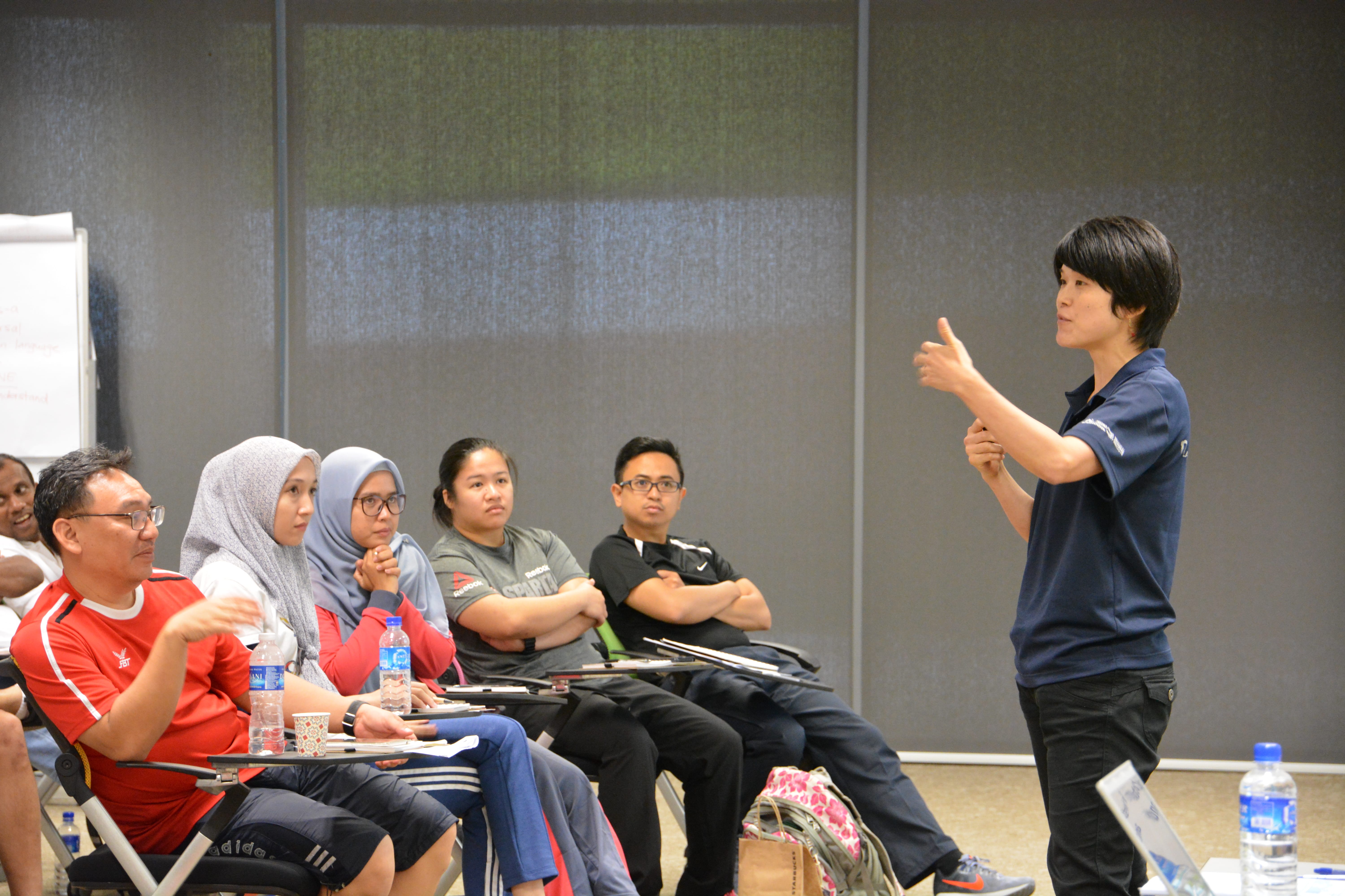 Presenter giving a thumbs-up to seated attendees in a classroom, with notebooks and water bottles on desks.