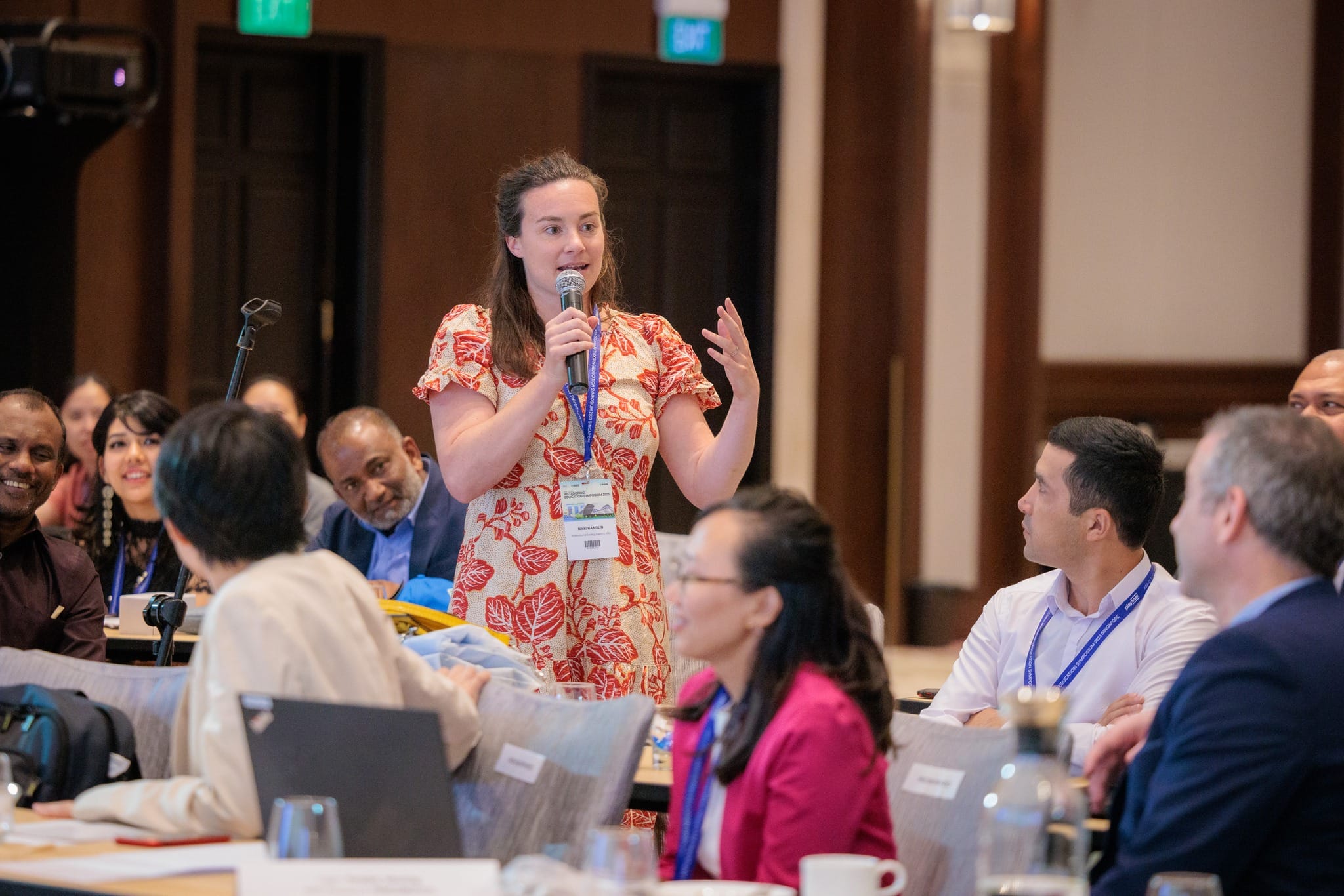 A woman in a red floral dress speaks into a microphone at a conference, surrounded by seated attendees wearing lanyards.