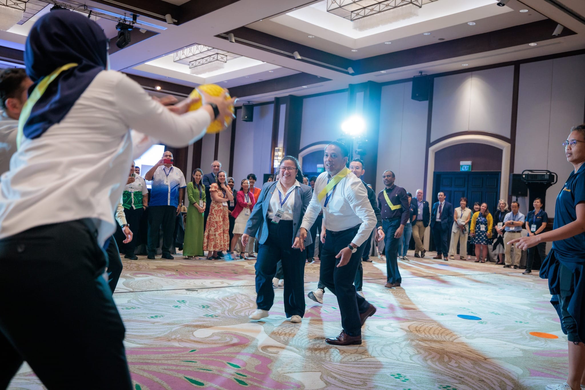 Two professionals play a ball game in a conference ballroom while a crowd of attendees watches and smiles.