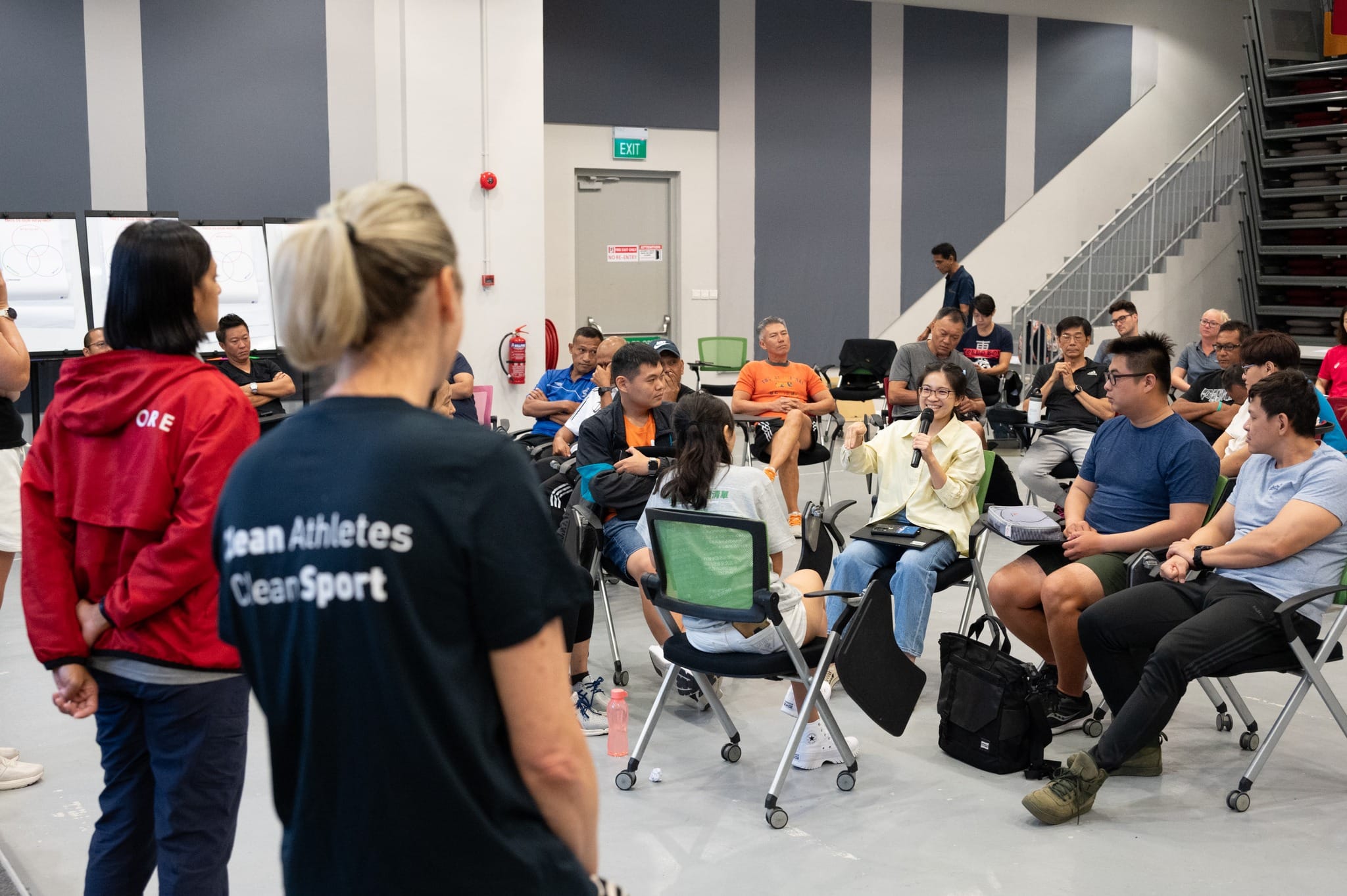 A group of people seated in chairs in a circle during a seminar, with a woman holding a microphone speaking.