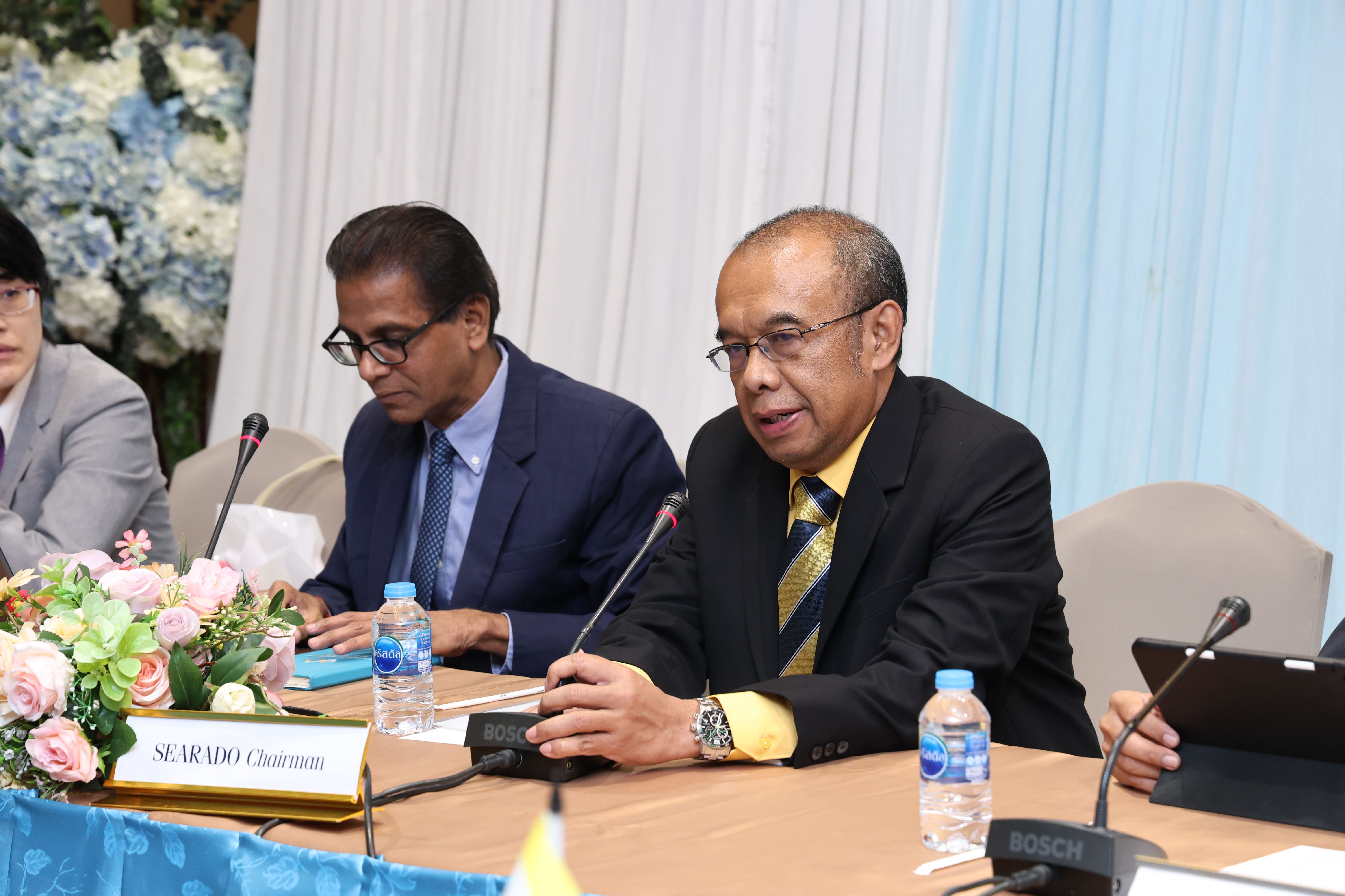 Man in black suit and yellow shirt speaks at a meeting table with microphones, water bottles, and a nameplate.