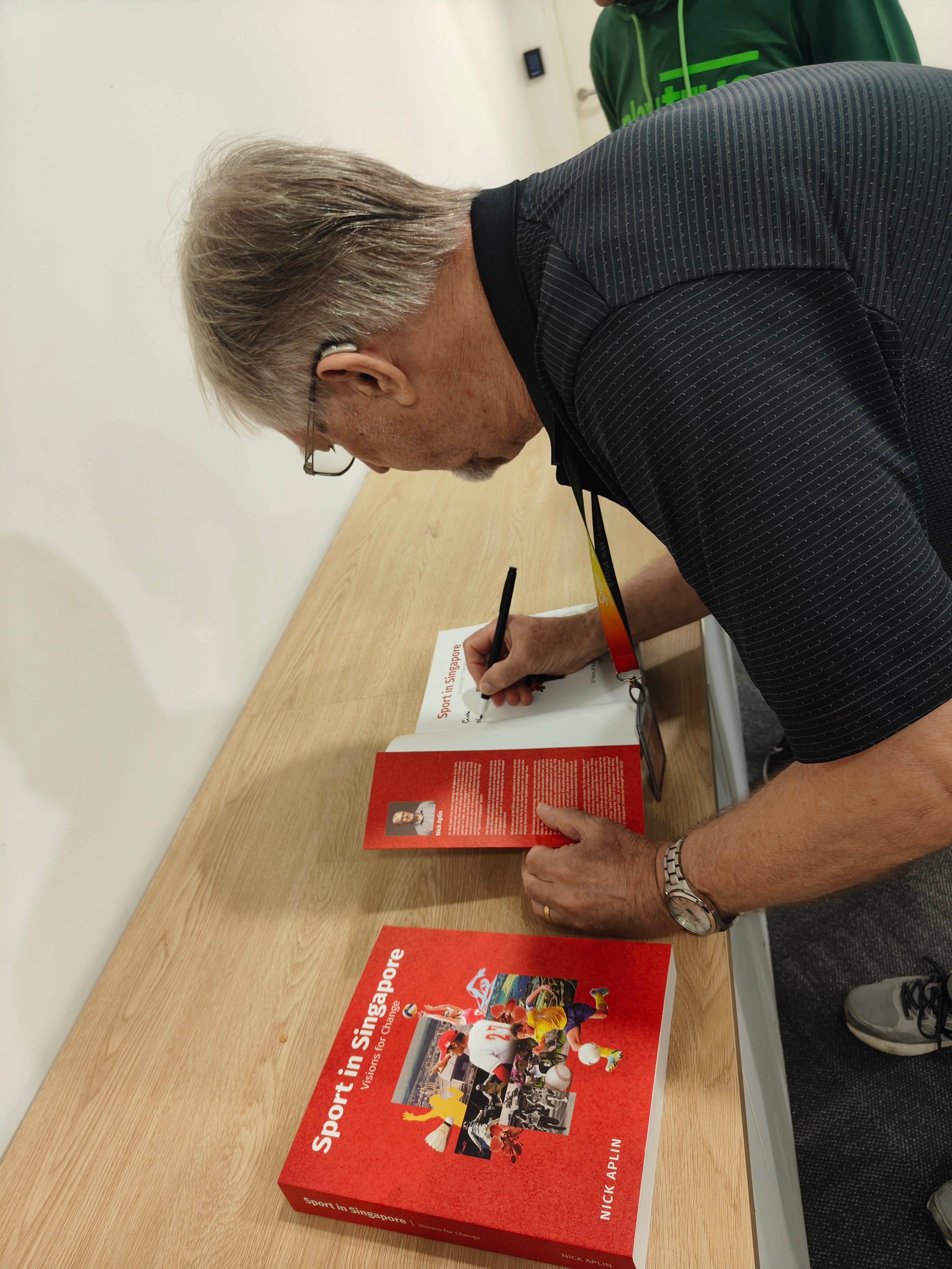 An older man with gray hair signs a red book titled Sport in Singapore by Nick Aplin on a wooden table.