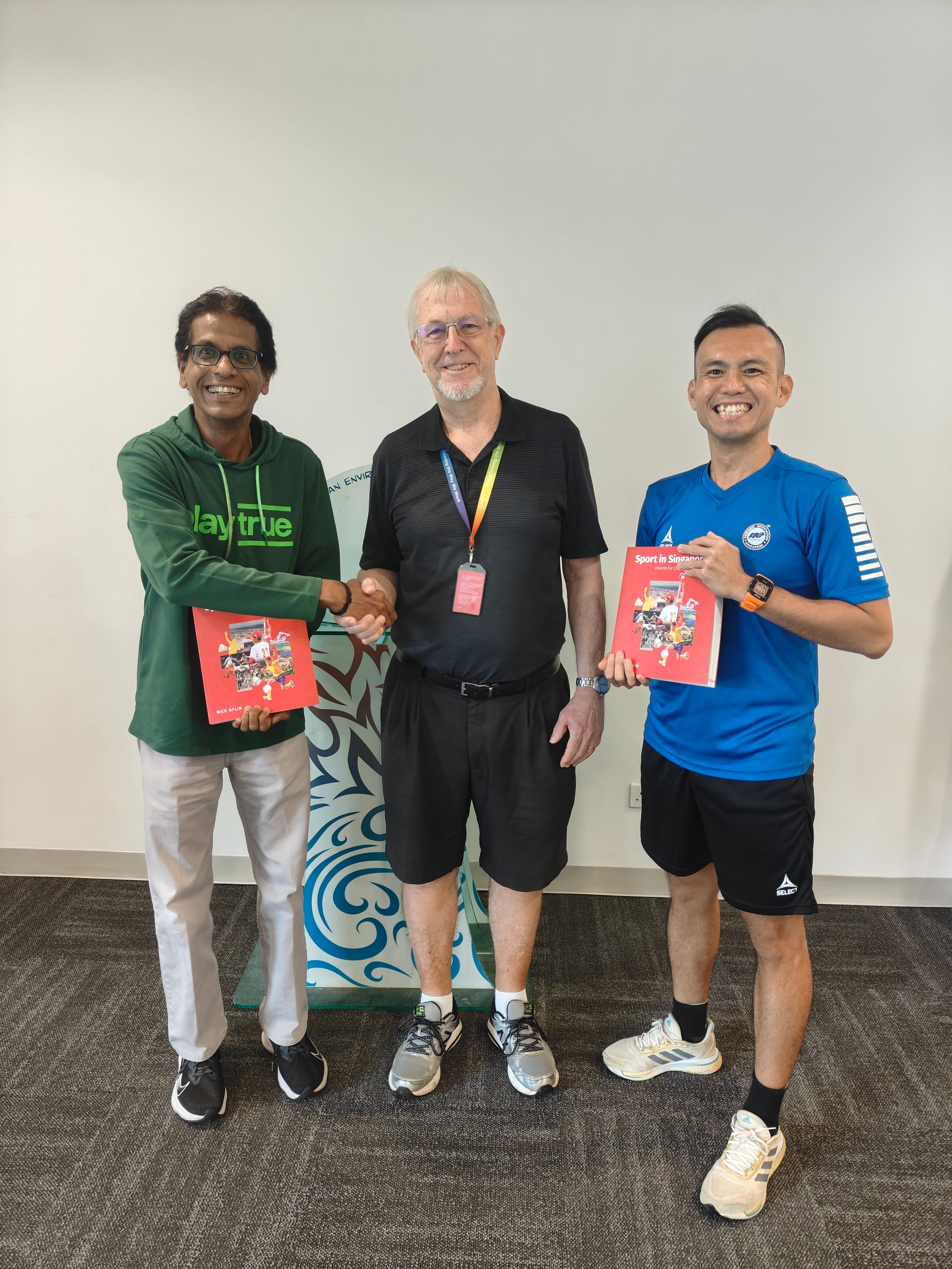 Three men standing indoors; left and center shake hands while left and right hold red books.