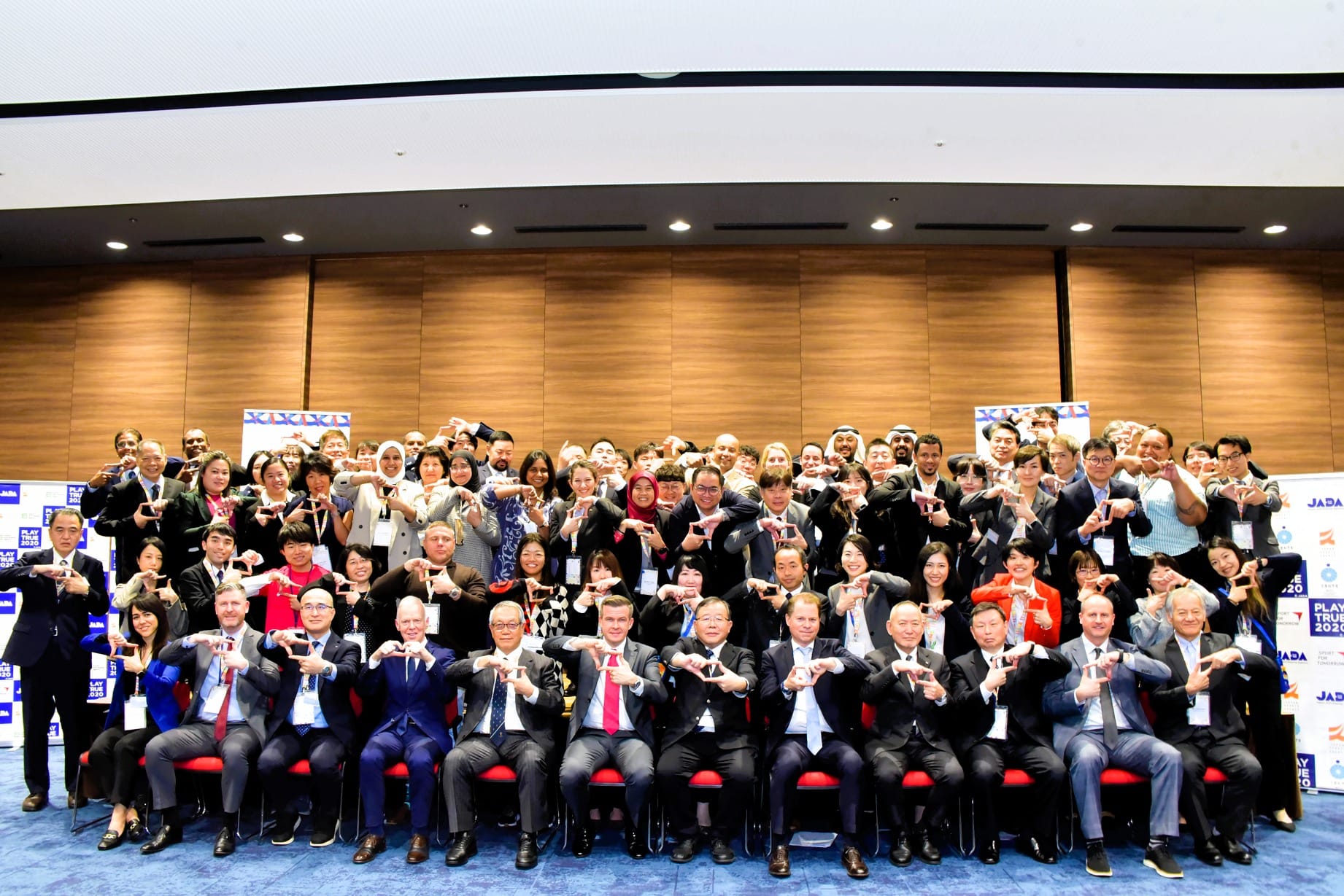 Large group of formally dressed conference attendees posing together, all making heart shapes with their hands.