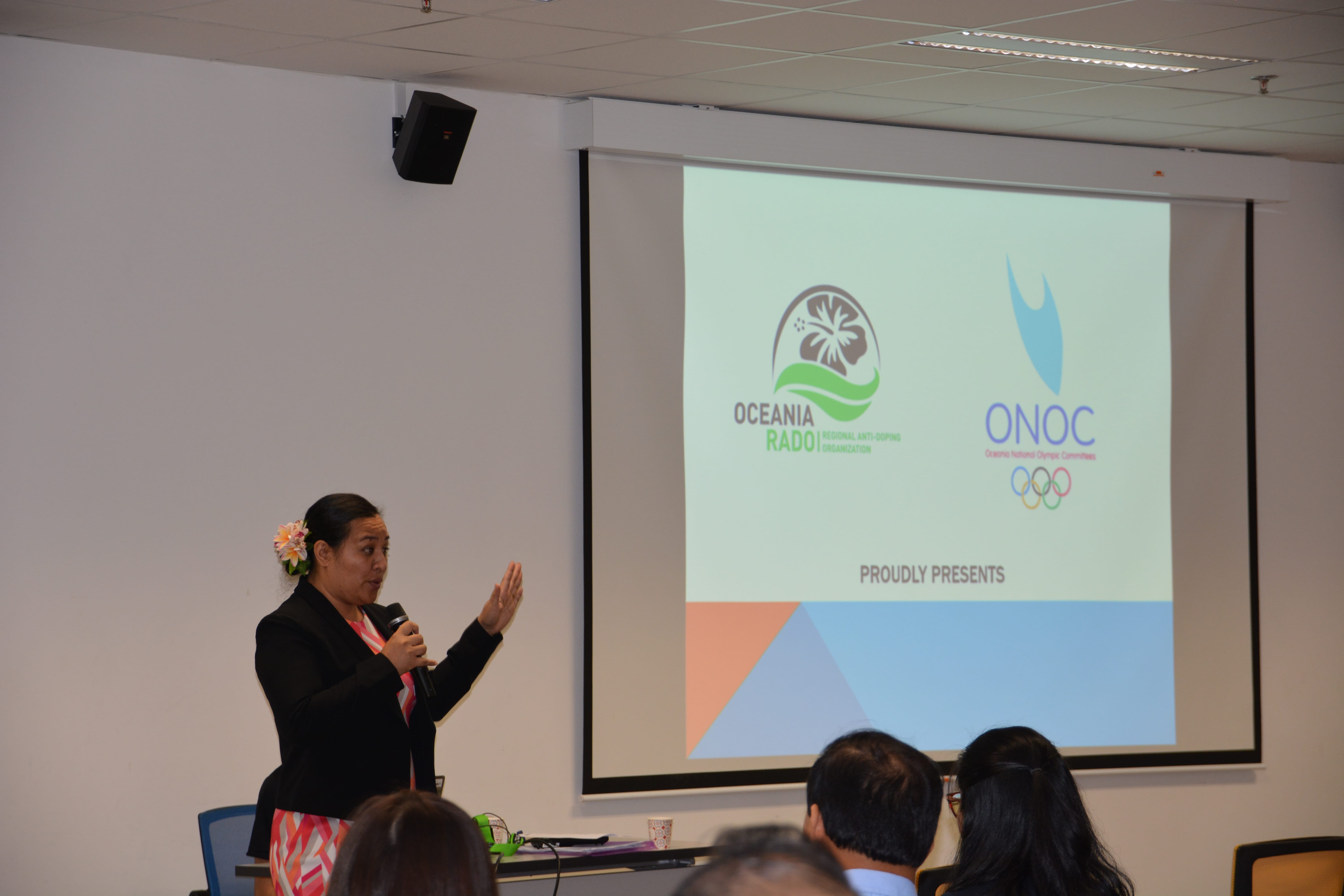 Speaker with microphone addresses seated audience beside projected slide with Oceania RADO and ONOC logos