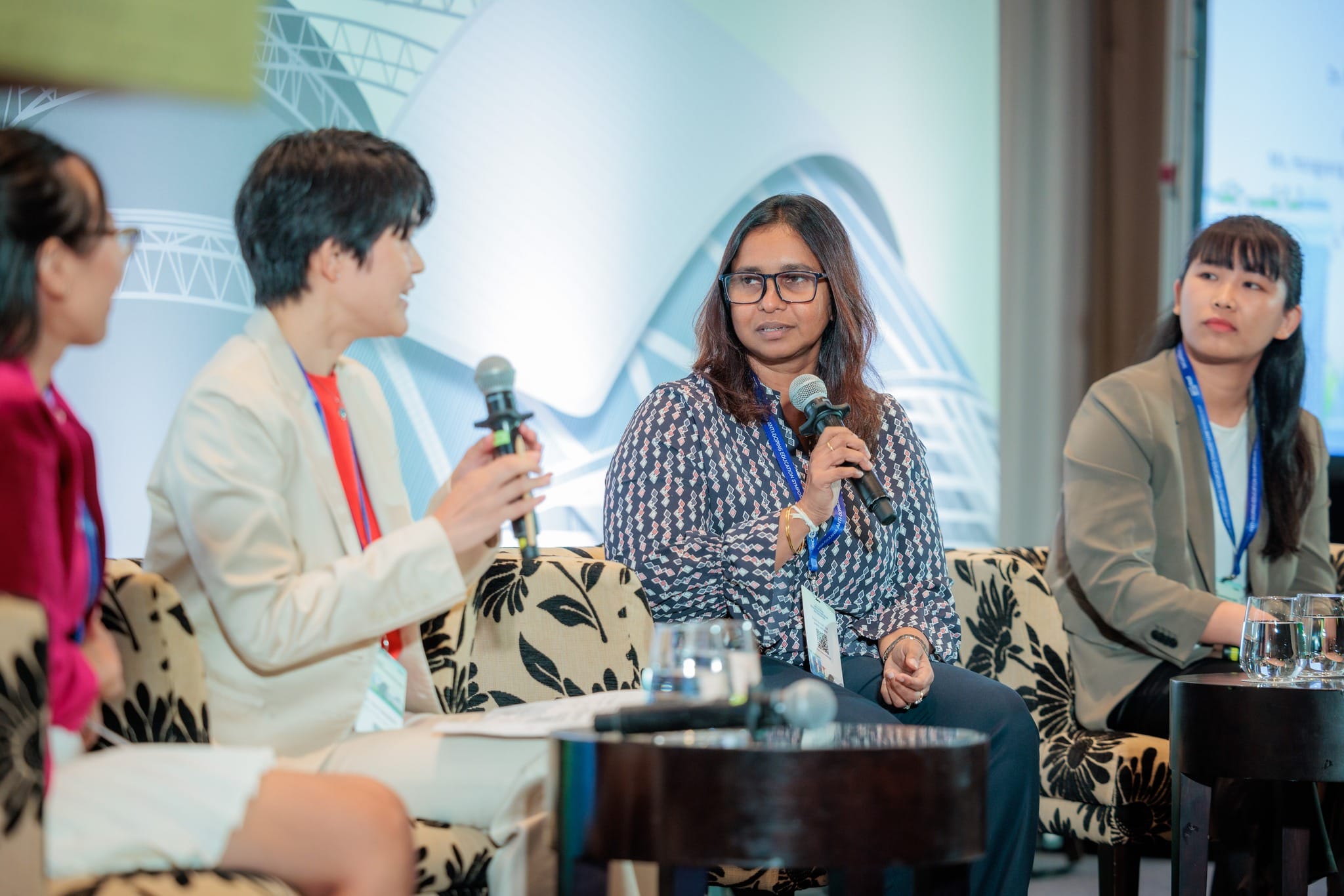 Four women sit on a panel discussion, two holding microphones and speaking, all wearing conference lanyards.