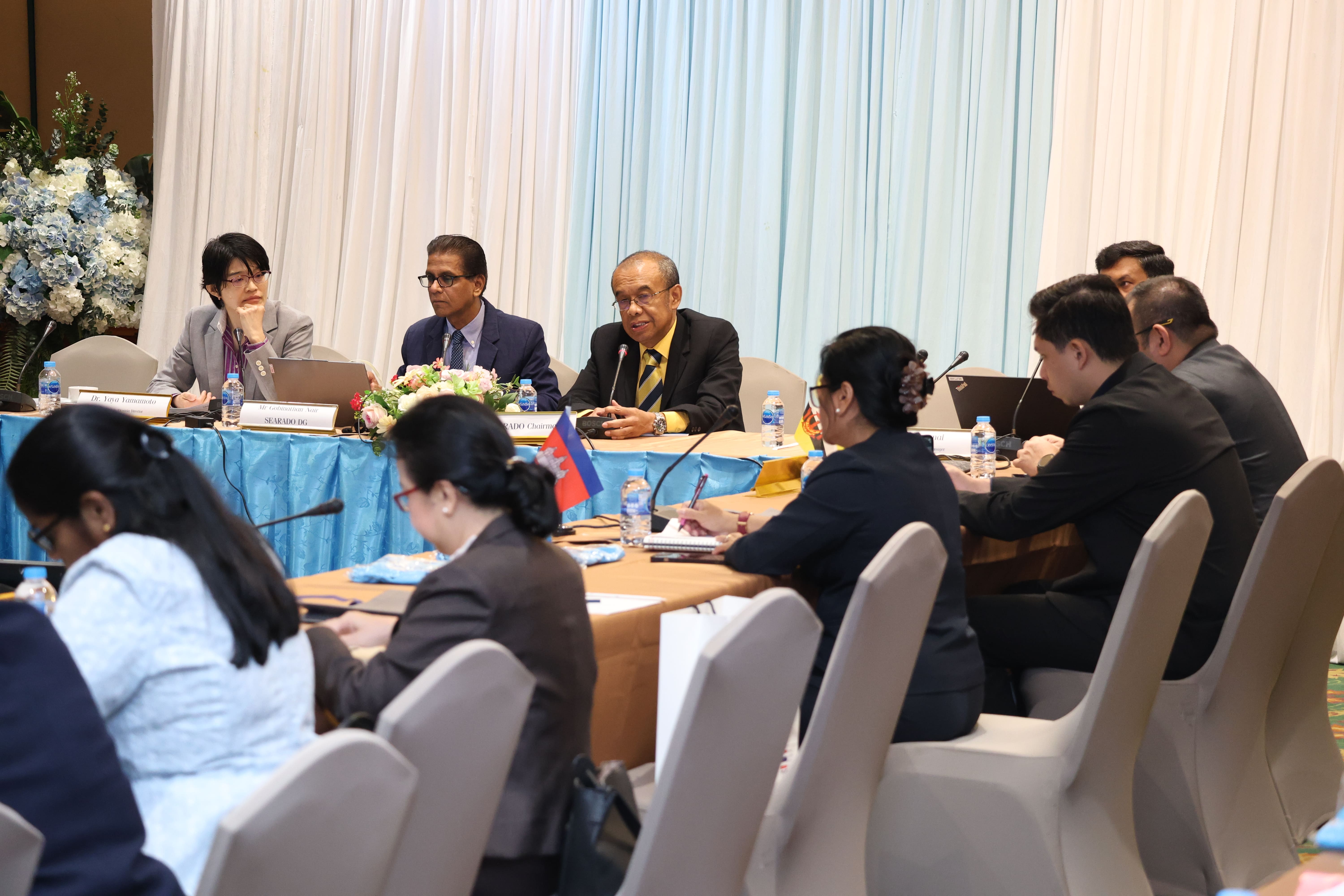 Panel discussion at a conference table with microphones, laptops, nameplates, and attendees taking notes.