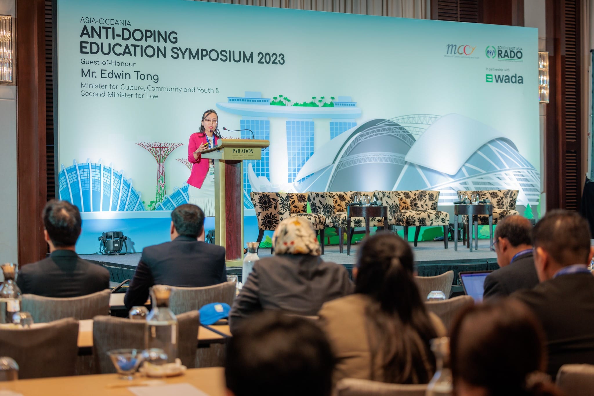 Woman in pink jacket speaks at podium during Asia-Oceania Anti-Doping Education Symposium 2023 before seated audience.