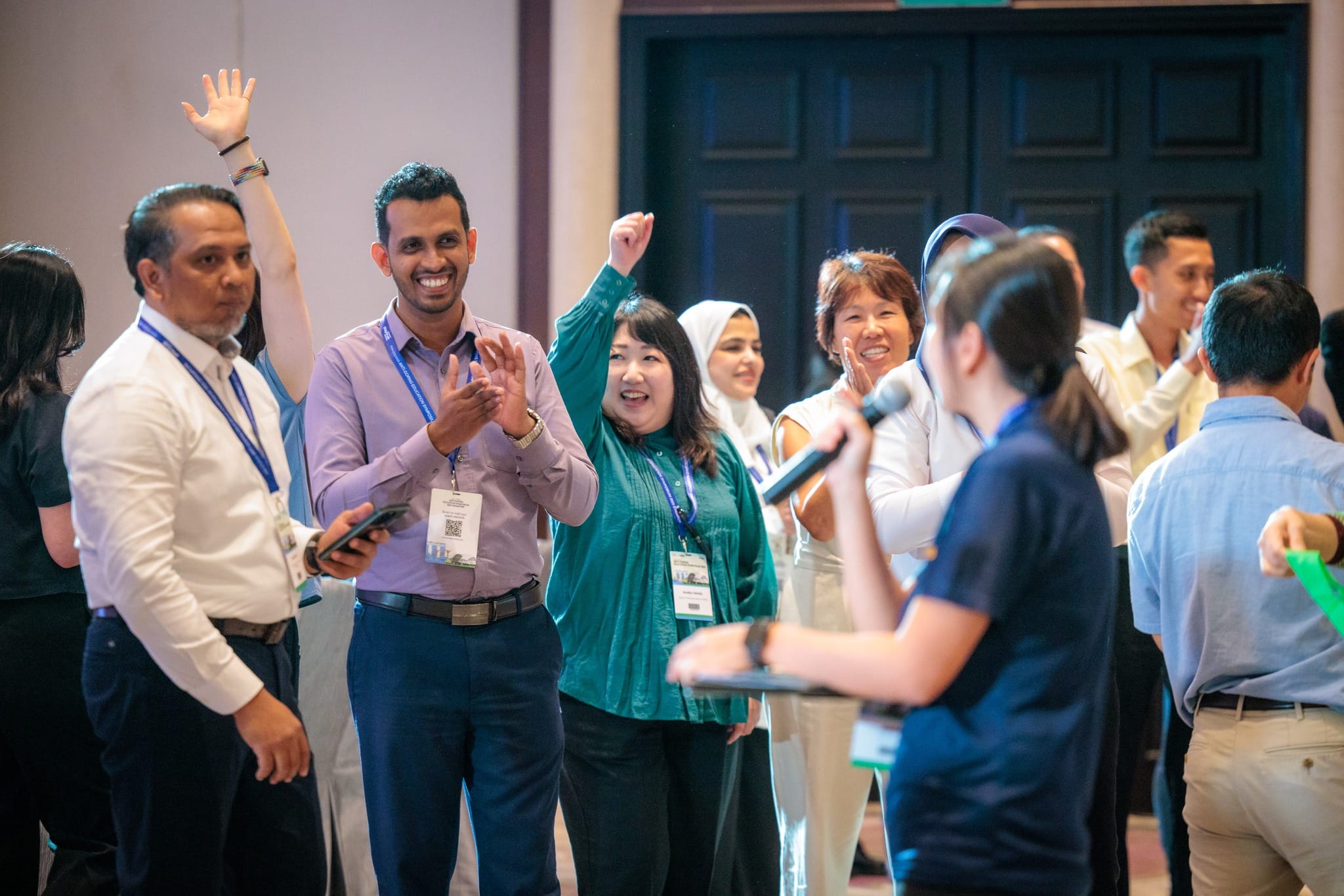 A group of smiling conference attendees with lanyards raise hands and clap while a person holds a microphone nearby.