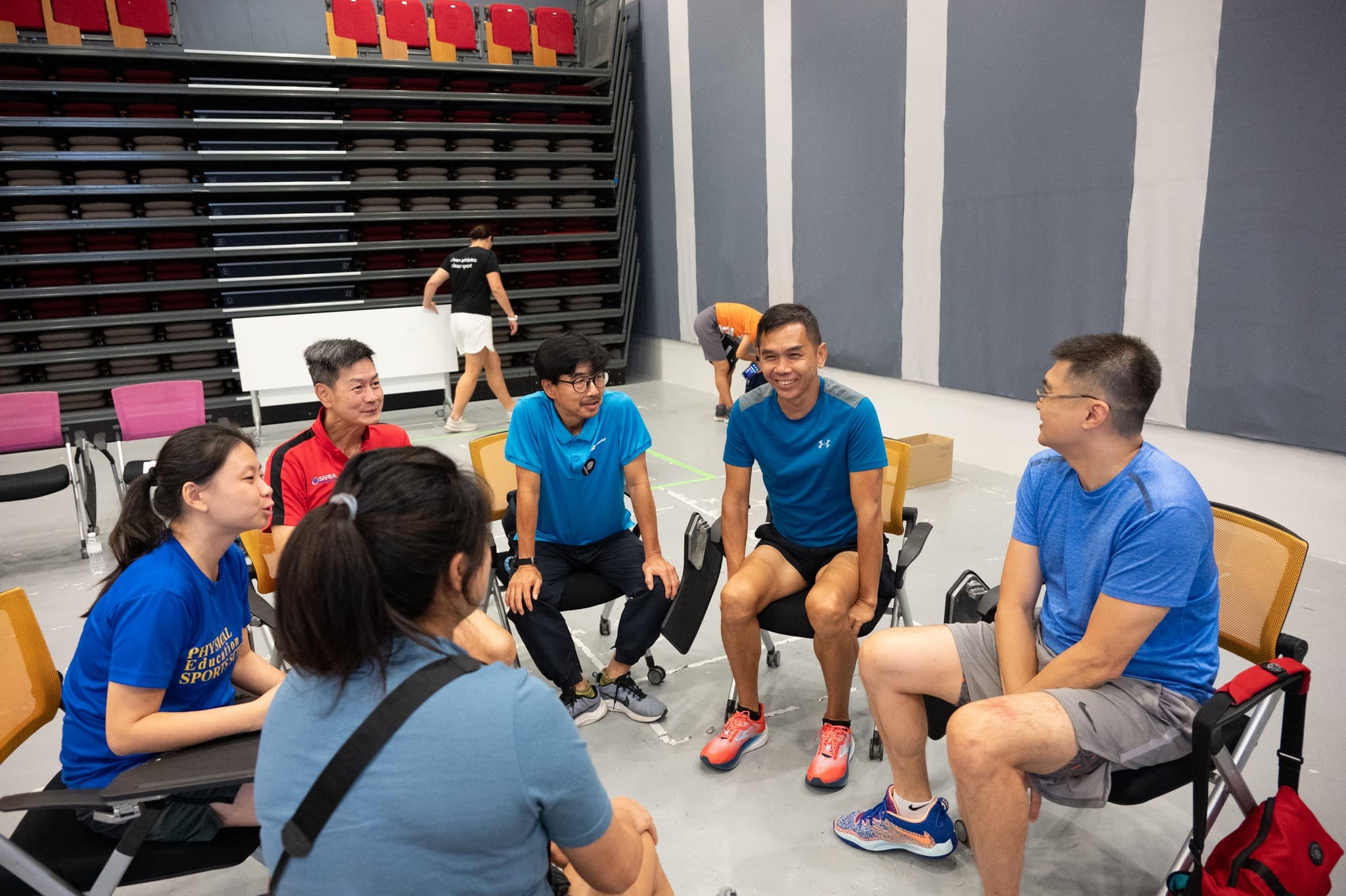 Five people in sportswear sit in a circle on chairs inside a gymnasium, chatting, with folded bleachers in the background.