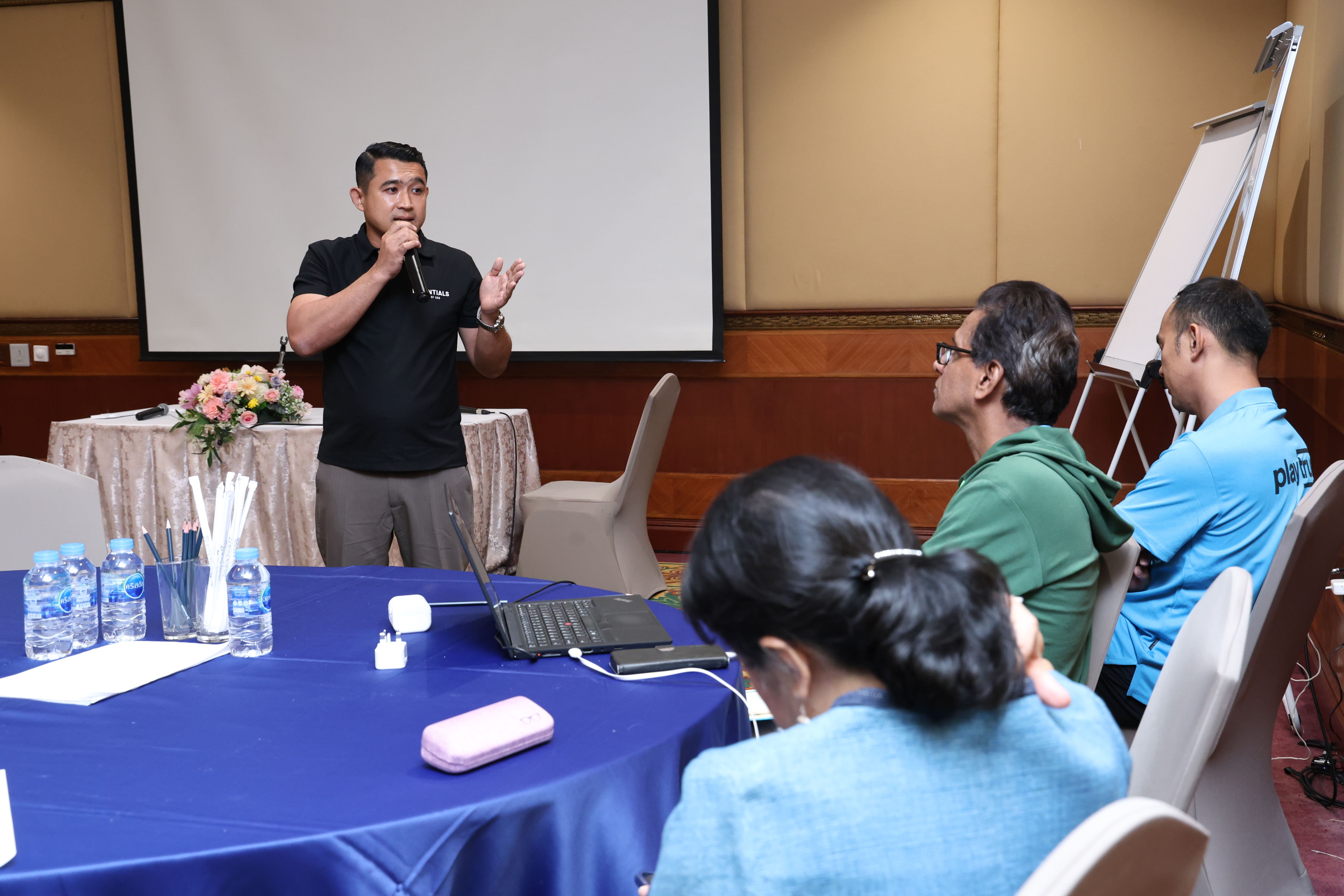 Man speaks into a microphone near a projector screen while seated attendees listen at a conference table.