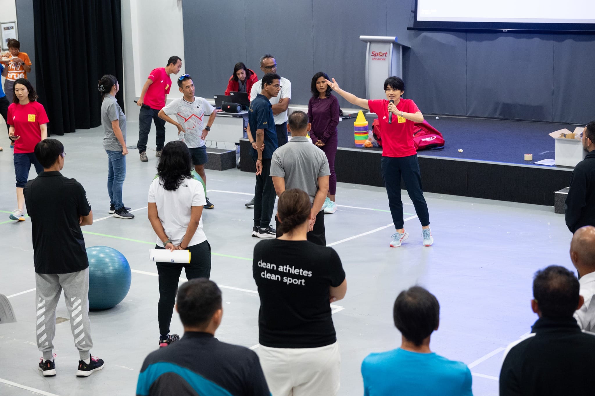A person in a red shirt holds a microphone and gestures while addressing a group in a Sport Singapore indoor hall.