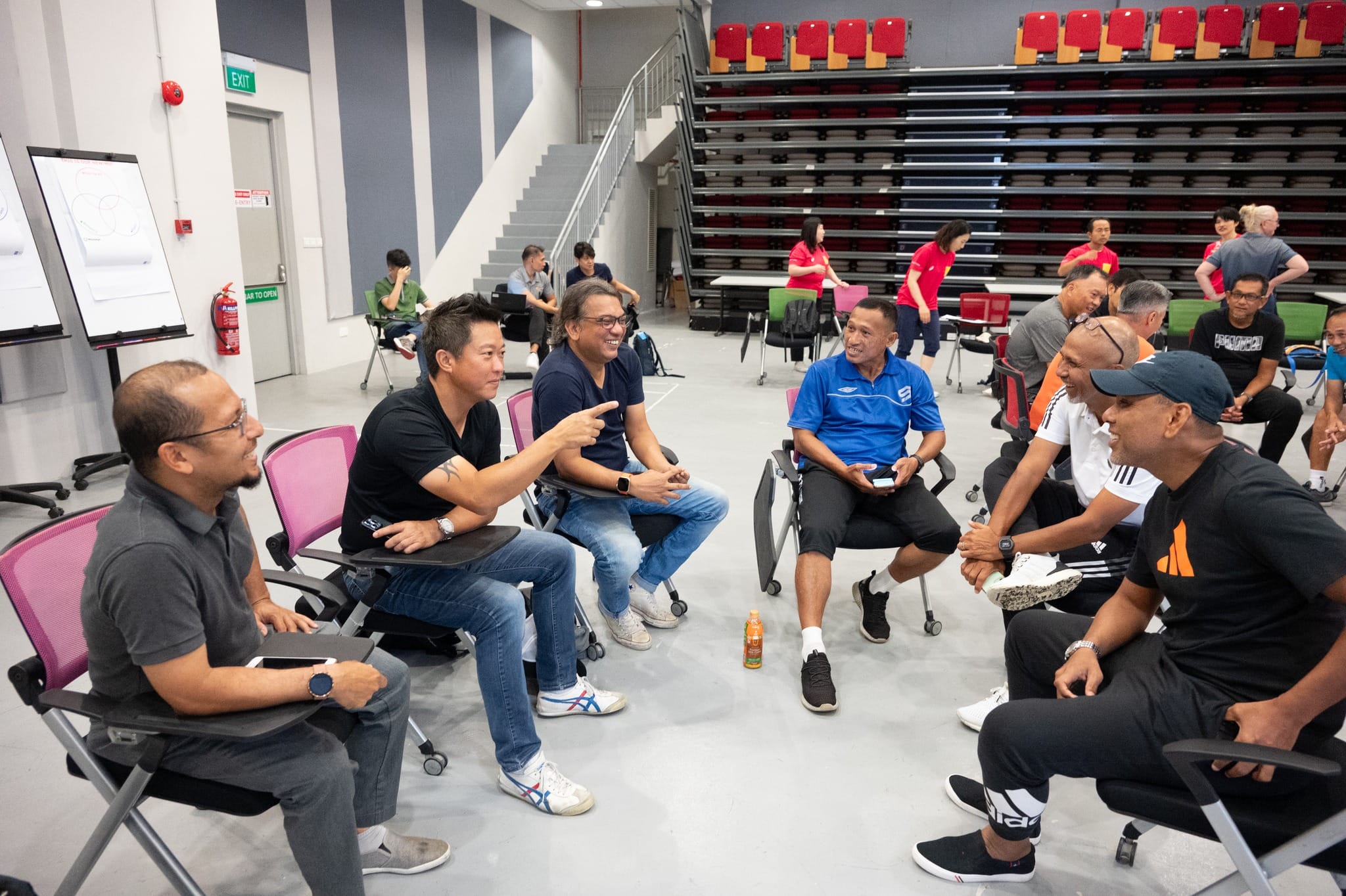 A group of men sit in a circle on chairs in a large indoor hall, smiling and talking during a workshop session.