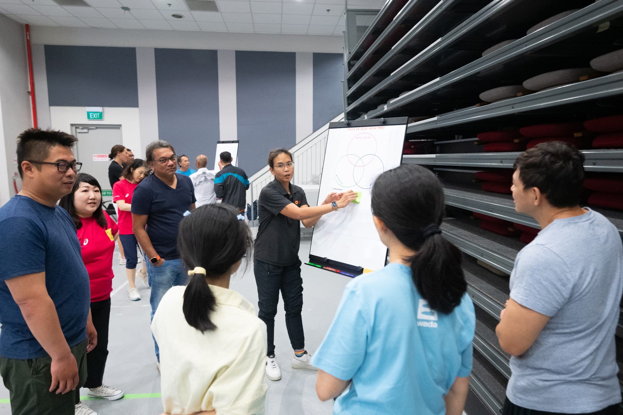 A woman in black points to a whiteboard with a Venn diagram while a group of people stands around her indoors.