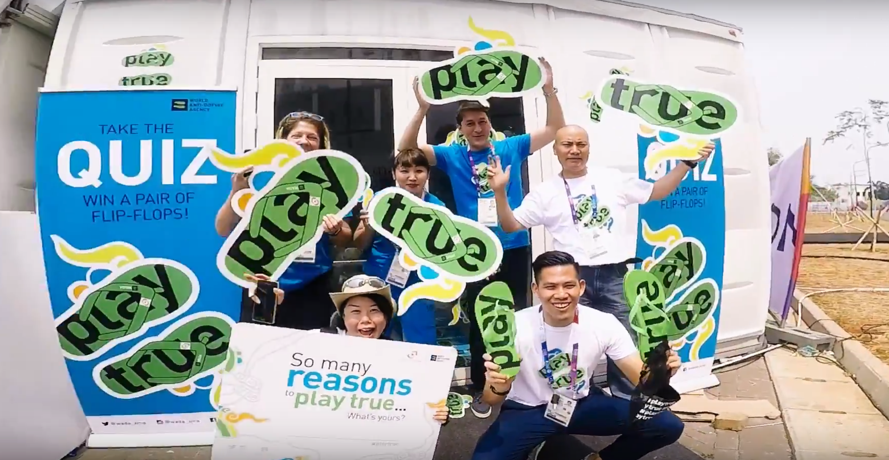 Six smiling people hold green flip-flop signs reading "play true" near a WADA quiz booth promoting clean sport.