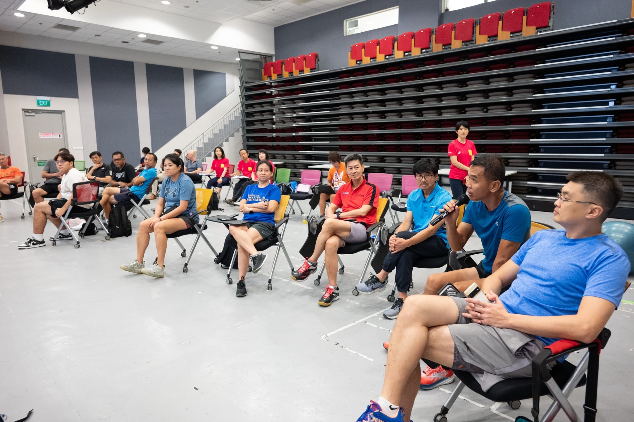 A group of people in athletic wear sit on chairs in a sports hall, with one man speaking into a microphone.