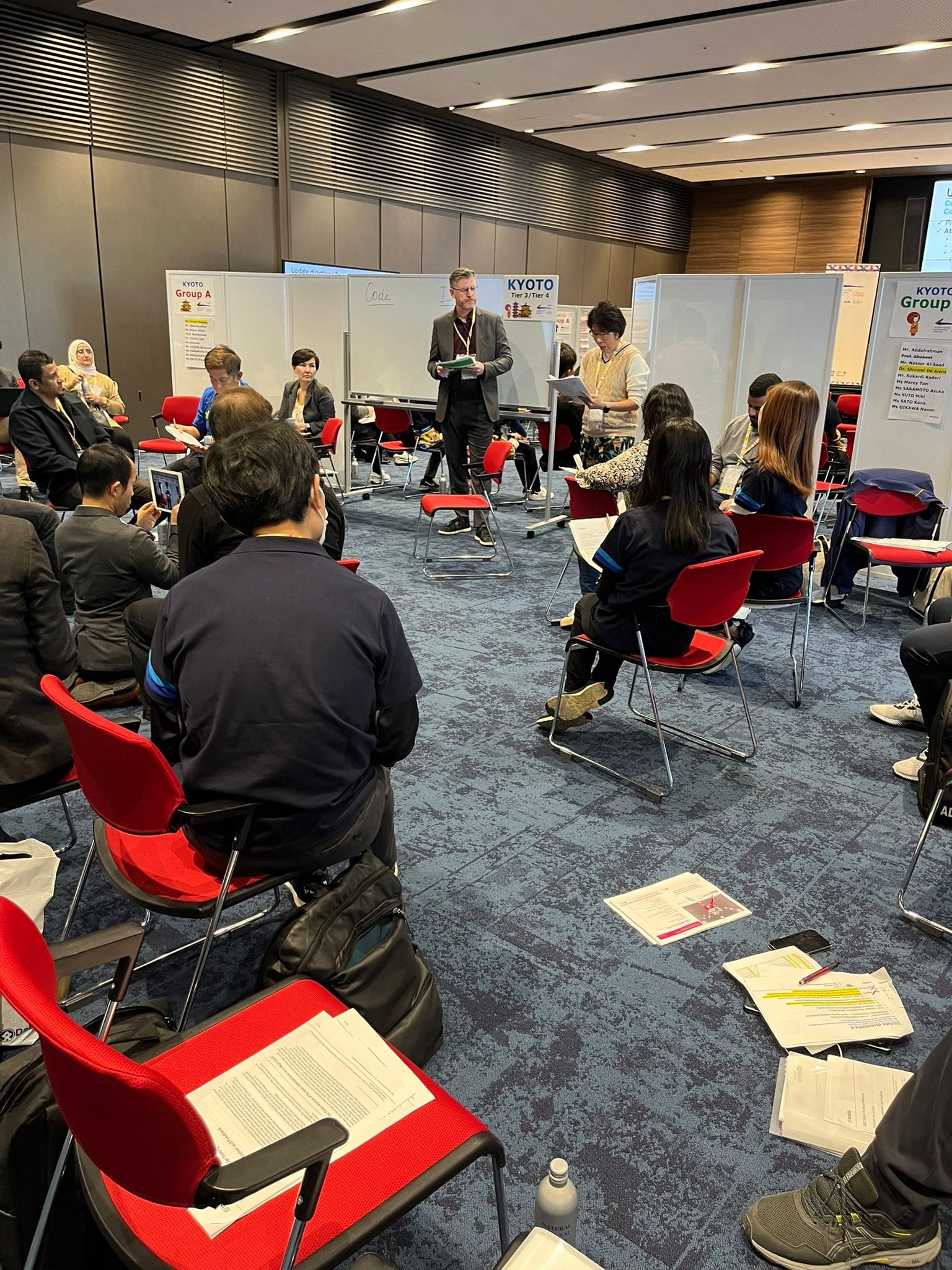 A man stands presenting to a seated group in a conference room with Kyoto Group A and Group signs on whiteboards.