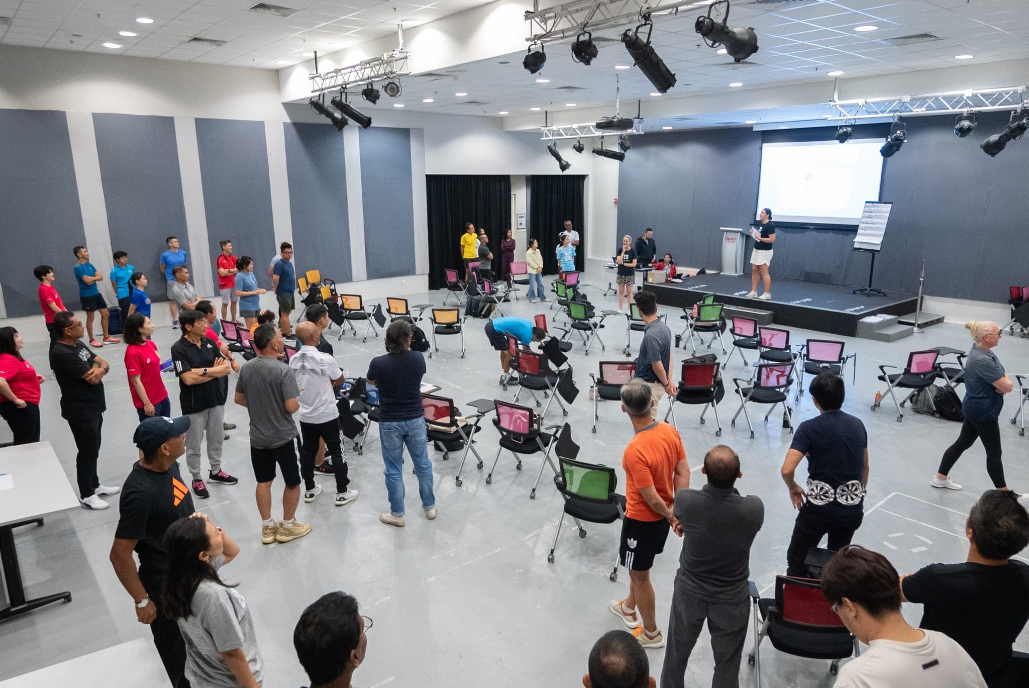 A large group of people stand in a circle in a hall while a presenter speaks near a projector screen.