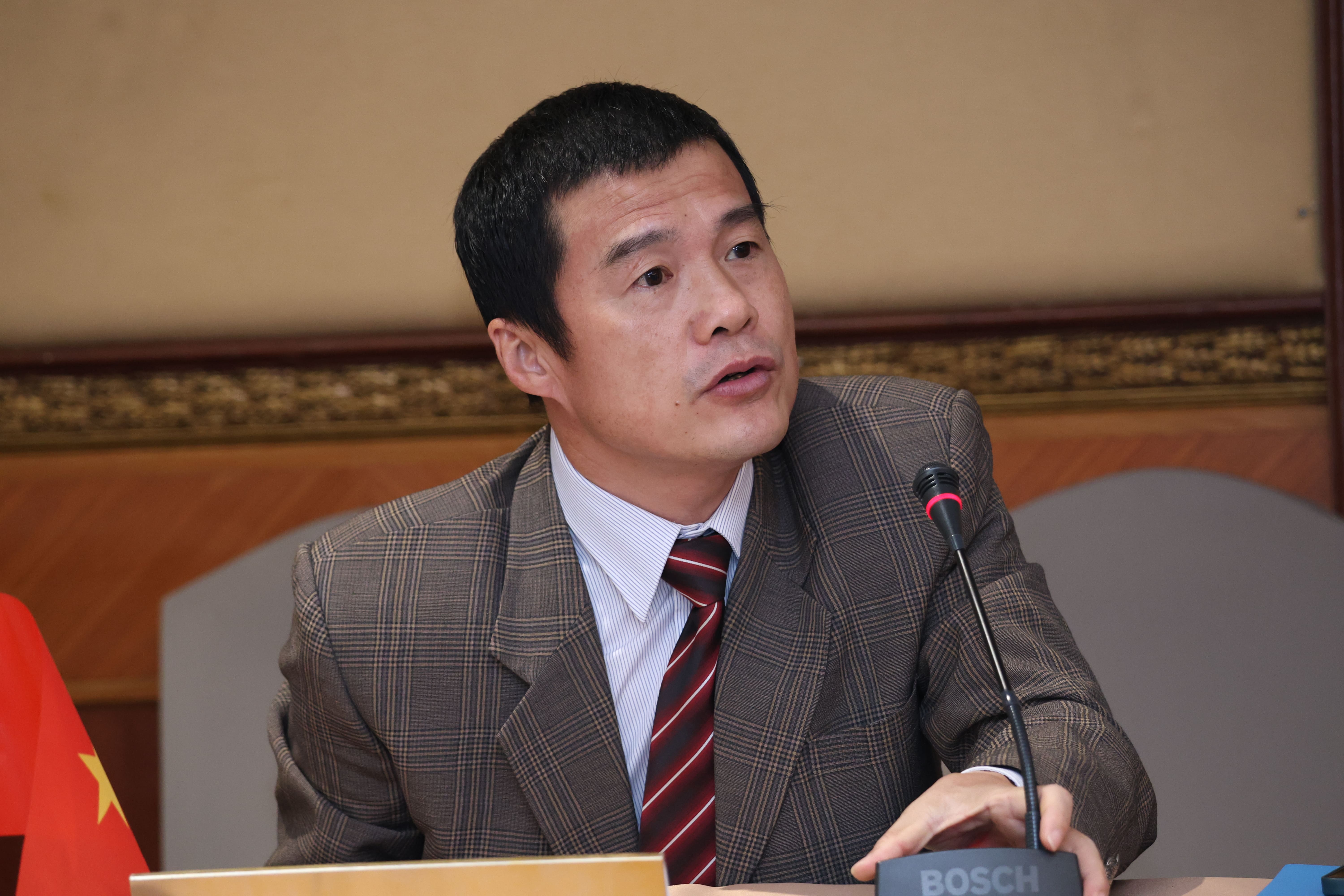 Man in plaid suit and striped tie speaks into a microphone at a table, looking to the side.