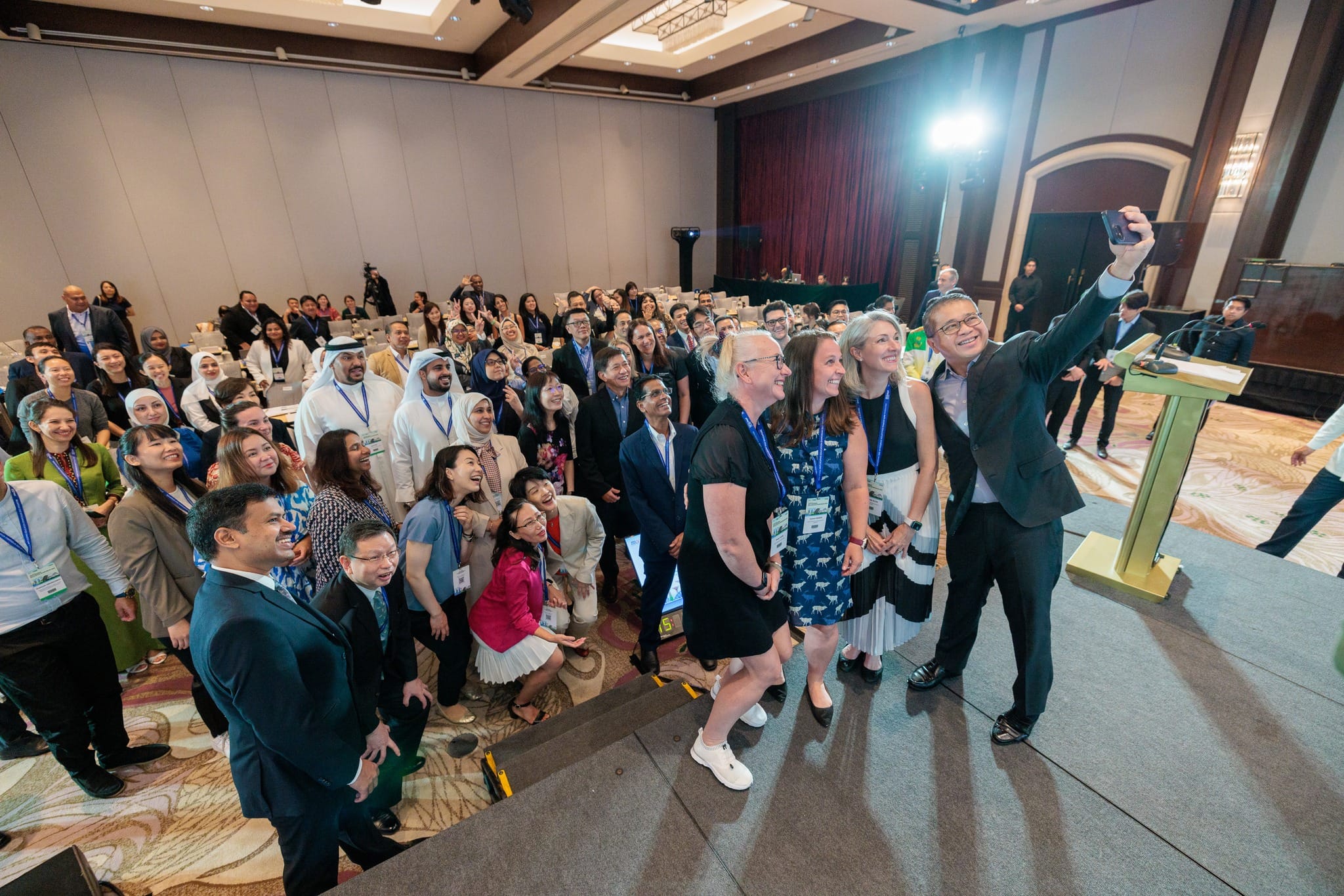 A large diverse group of conference attendees smiles as a man on stage takes a group selfie with his phone.