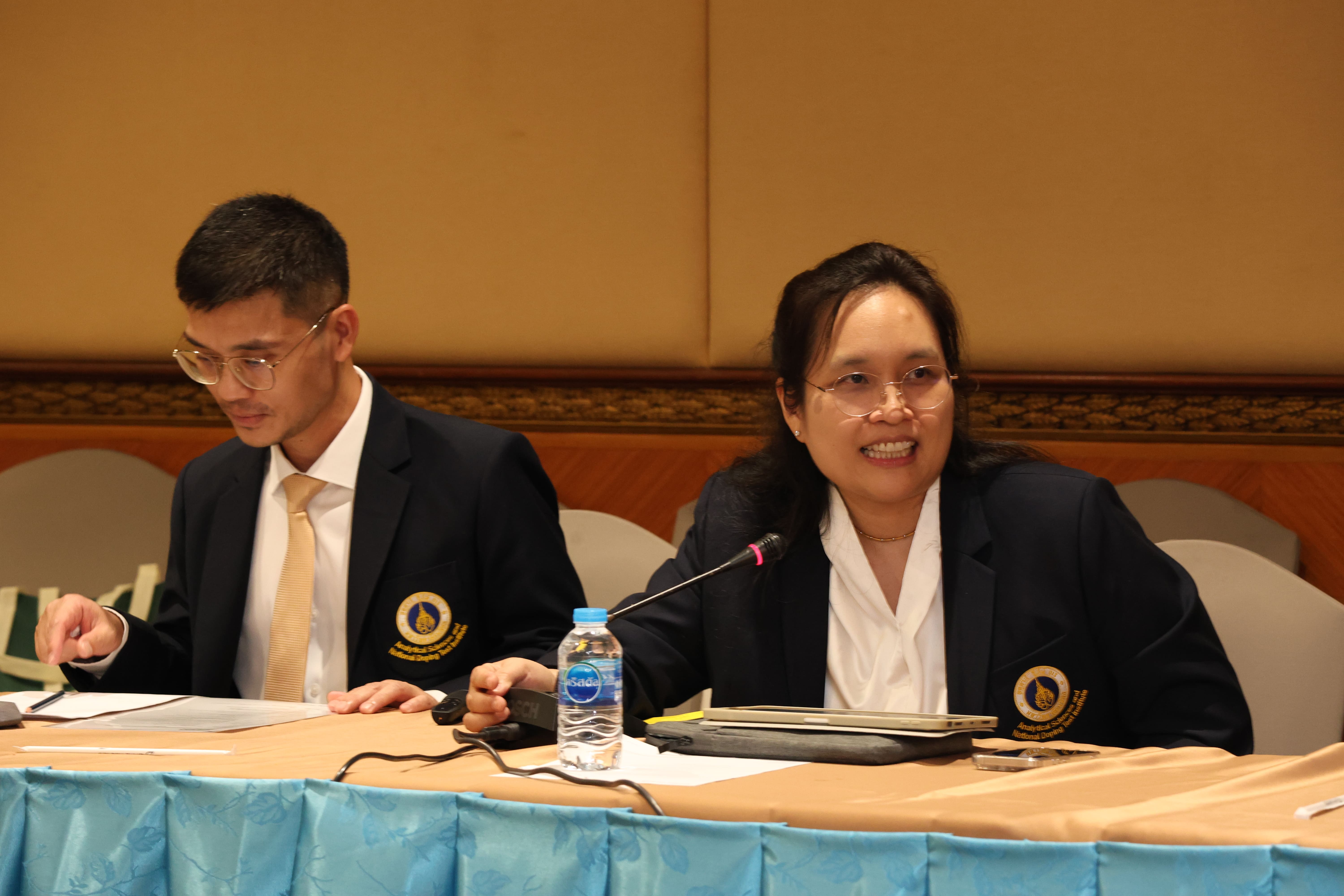 Two people in navy blazers sit at a conference table, one speaking into a microphone beside a water bottle.