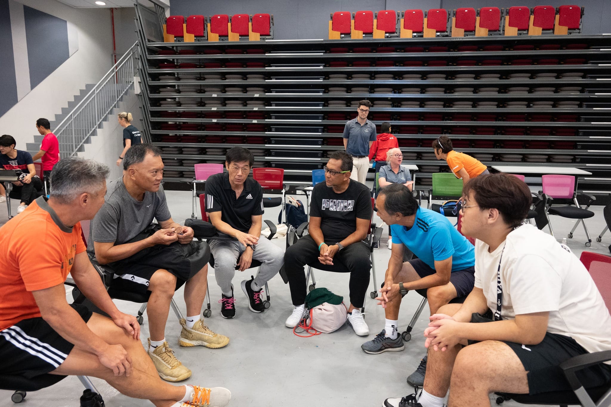 Six men in sportswear sit in a circle on chairs inside a sports hall with empty red bleachers behind them.
