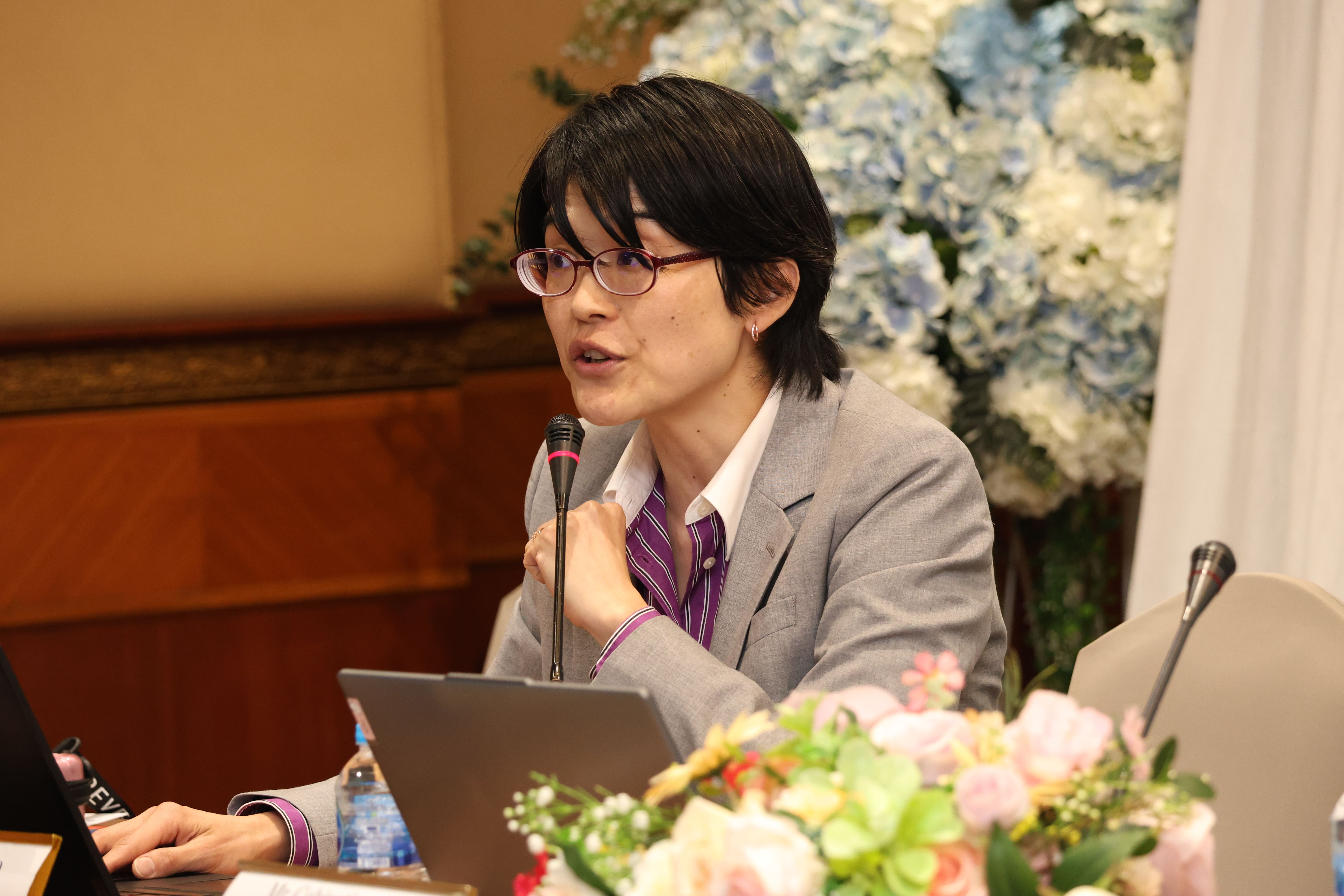 Person in glasses and gray blazer speaking into a microphone at a table with a laptop and flowers behind.